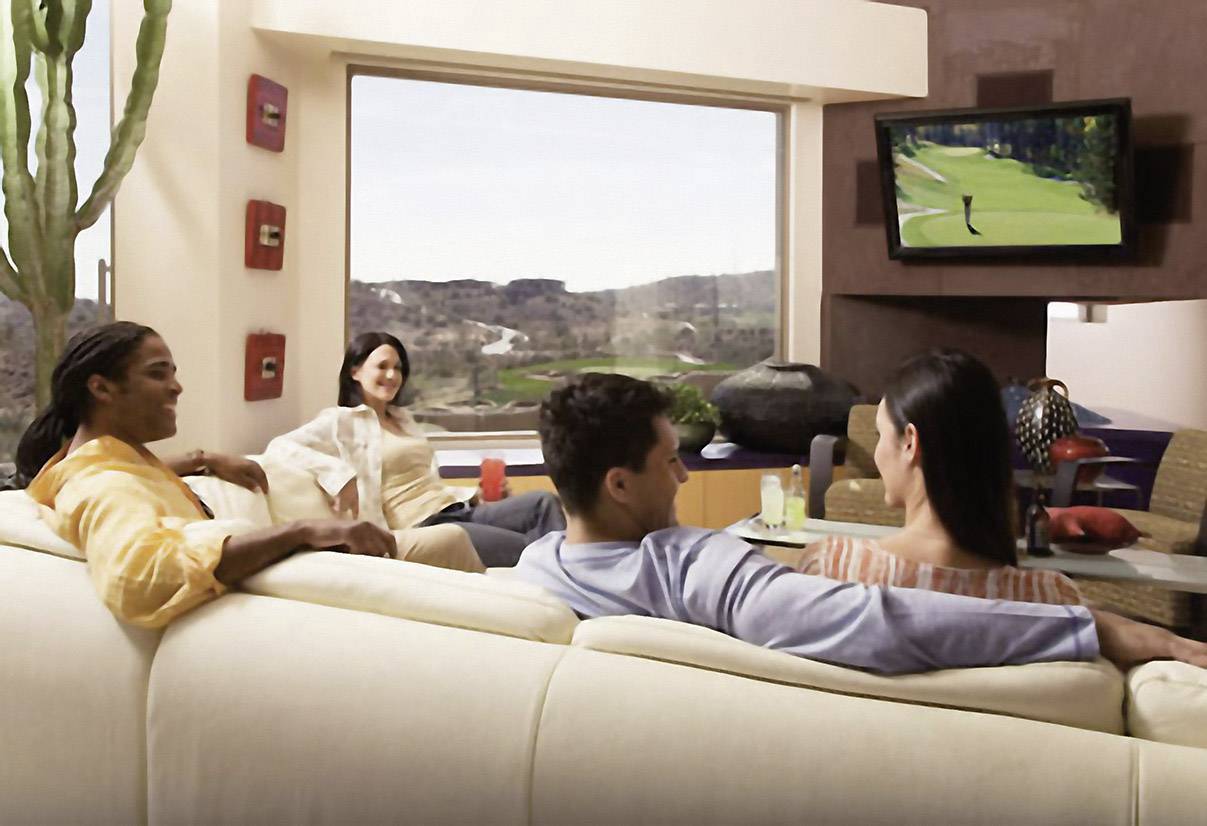 Cztery osoby siedzą swobodnie na kanapie i oglądają golfa w telewizji. Duży kaktus stoi w rogu pokoju.