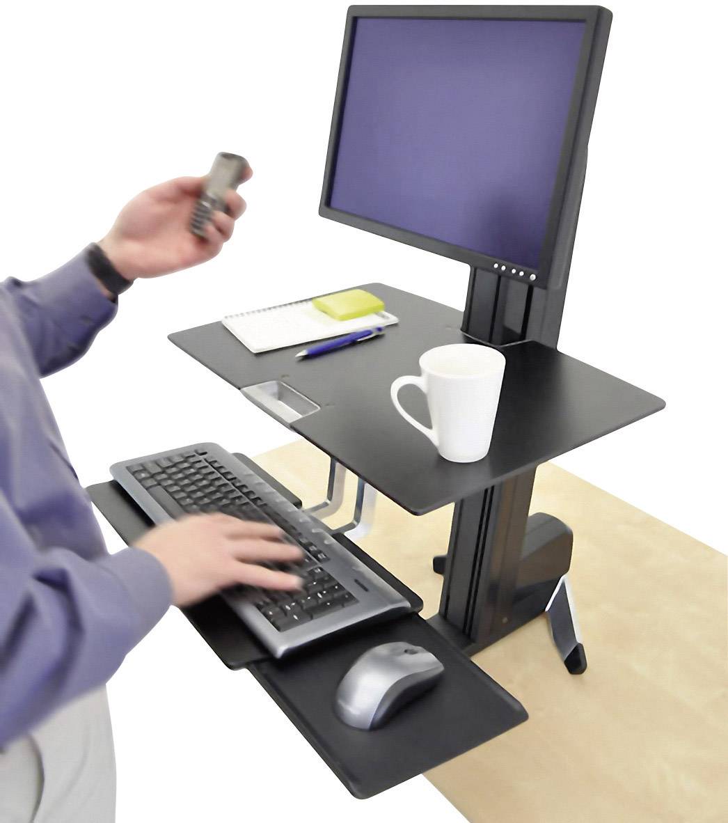 Osoba pracuje przy regulowanym biurku stojącym z monitorem, klawiaturą i myszą. Na blacie stoją kubek i notes.