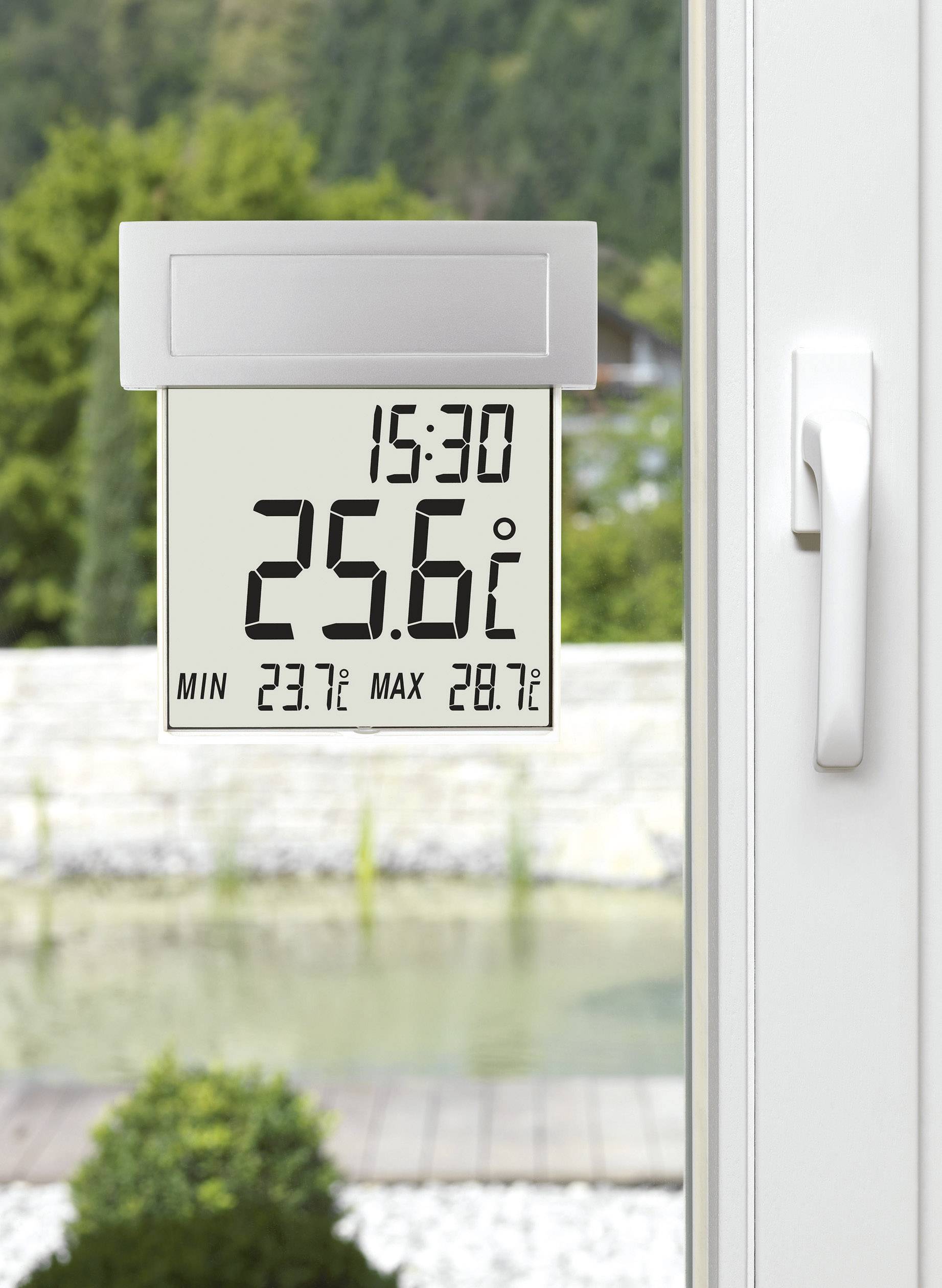 Cyfrowy termometr przy oknie wskazuje godzinę 15:30 i temperaturę 25,6°C, z minimum 23,7°C i maksimum 28,7°C. W tle: ogród.