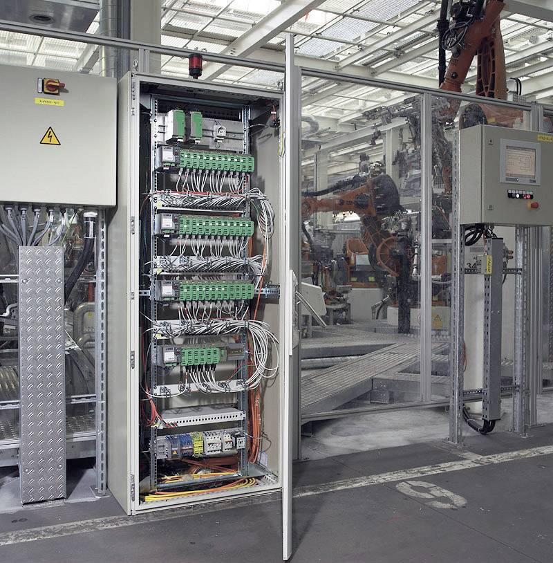 Otwarty szafa sterownicza ukazuje różnorodność kabli i elementów elektrycznych. W tle widoczna jest linia produkcyjna.