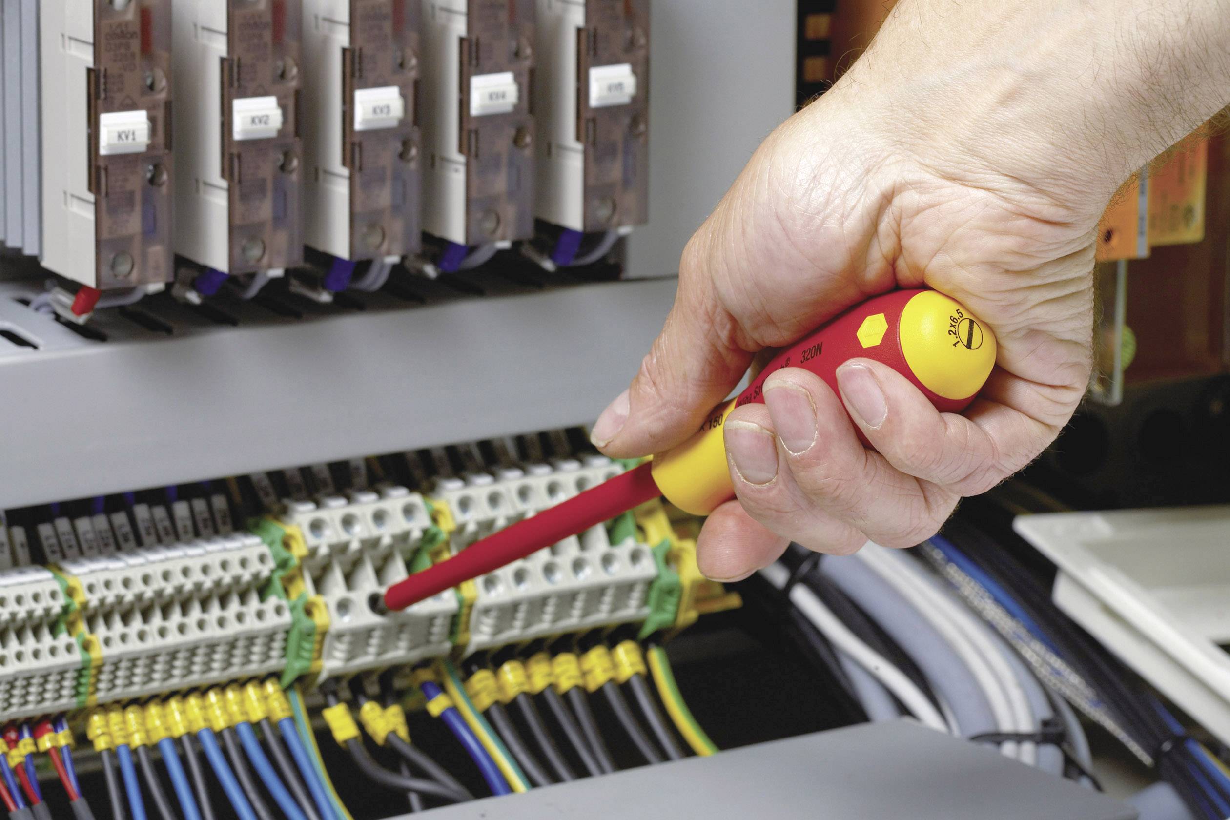 Jedna ręka trzyma śrubokręt i pracuje przy otwartej szafce elektrycznej, otoczonej przewodami i podłączeniami.