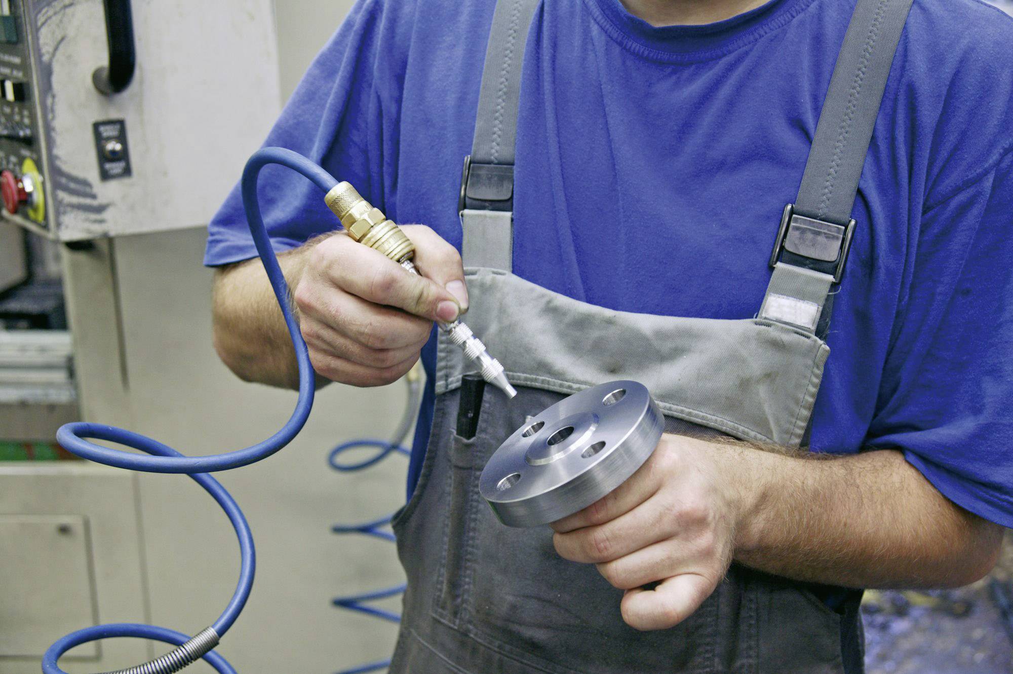 Robotnik w niebieskim kombinezonie oczyszcza część metalową sprężonym powietrzem w warsztacie.