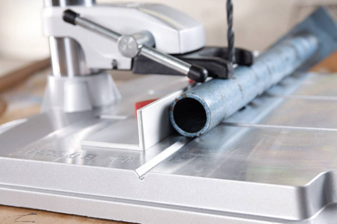 Stojak wiertarski służy do precyzyjnej obróbki rury metalowej. Niebieska rura jest mocno zaciśnięta pod wiertłem.