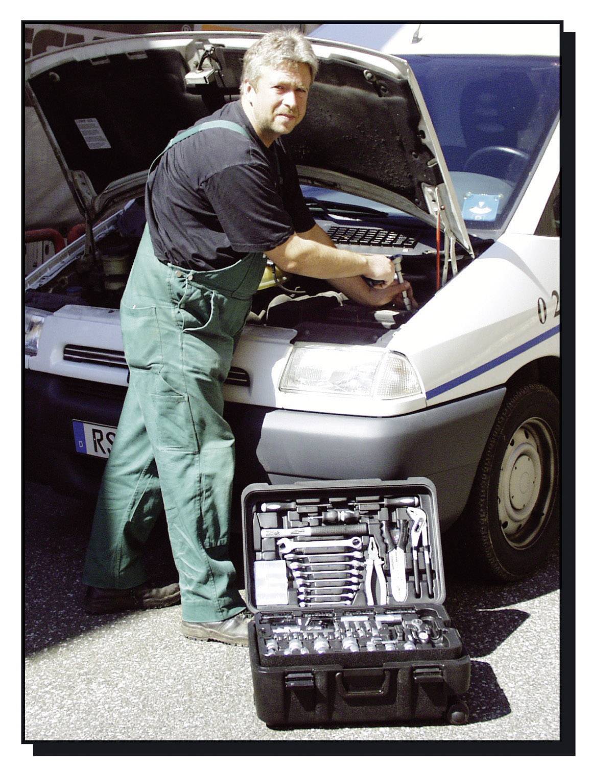 Mechanik w zielonym kombinezonie naprawia silnik białego pojazdu. Przed nim na podłodze stoi otwarty skrzynka narzędziowa.