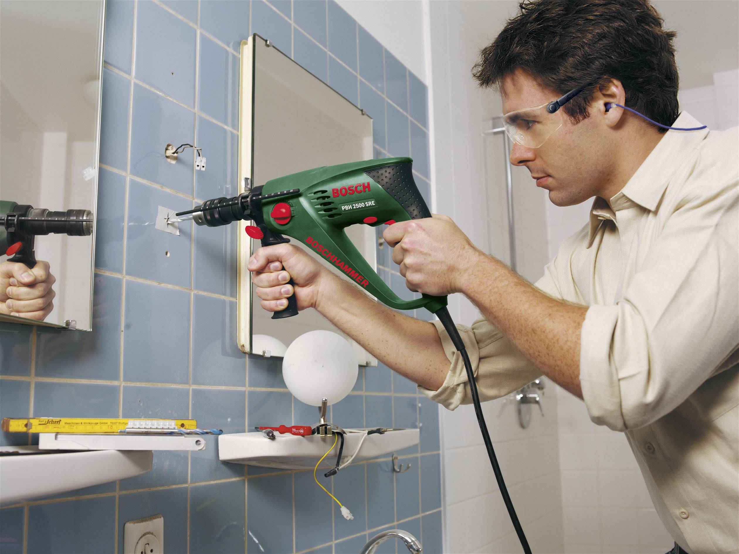 Mężczyzna w okularach ochronnych używa wiertarki, aby wywiercić otwór w wykafelkowanej ścianie łazienki. Narzędzia leżą w pobliżu.
