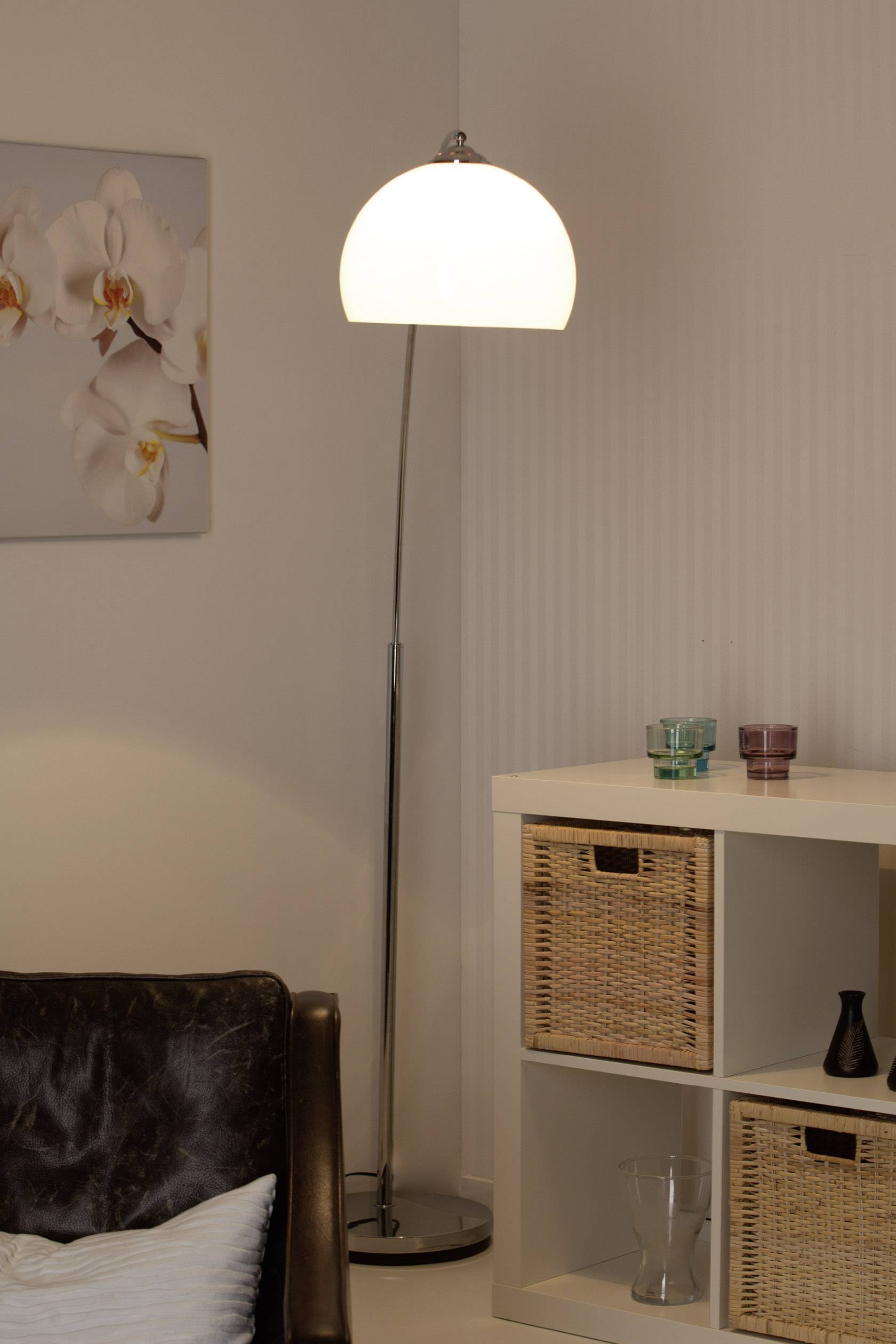 Lampa stojąca świeci obok regału z koszami w nowoczesnym salonie. Na ścianie w tle wisi obraz z kwiatami.