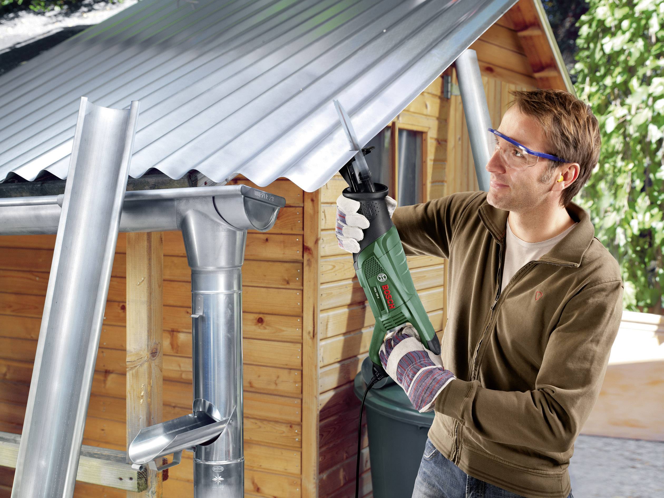 Mężczyzna piłą elektryczną nacina dach z blachy falistej na drewnianym domku. Ma na sobie okulary ochronne i rękawice robocze.