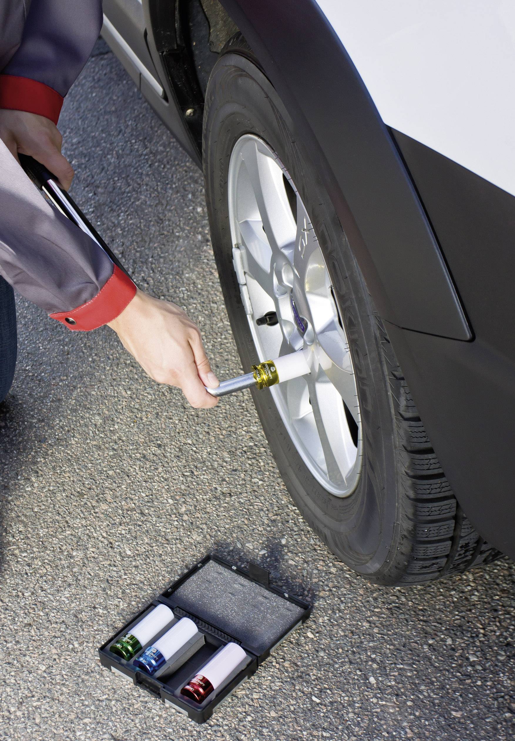 Osoba wymienia oponę samochodową kluczem, ujęcie z bliska. Skrzynka narzędziowa na ziemi z kolorowymi nasadkami.