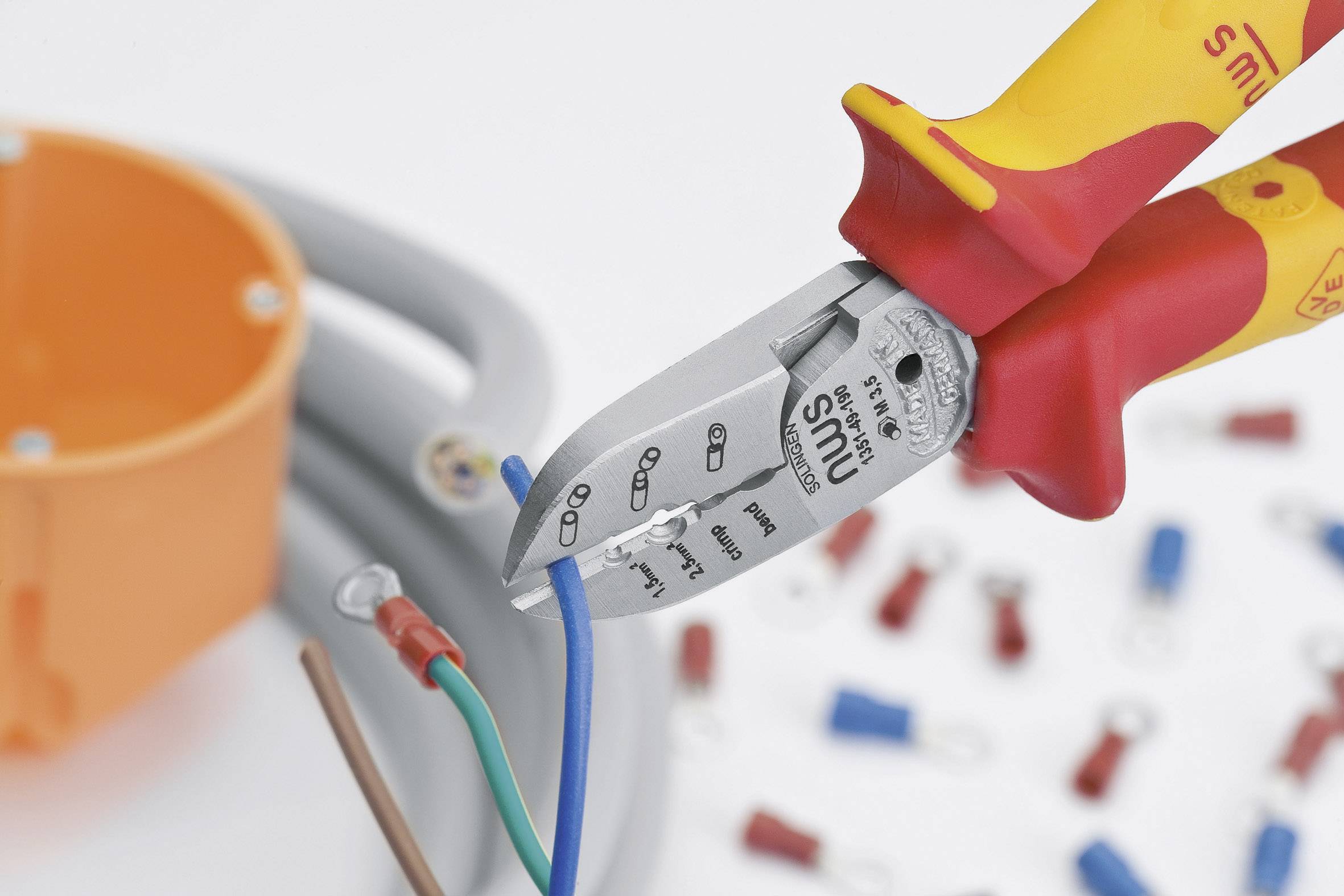 Szczypce elektryka przecinają niebieski kabel obok szarej szpuli przewodu i pomarańczowego pudełka z tworzywa sztucznego. W tle leżą kolorowe końcówki kablowe.