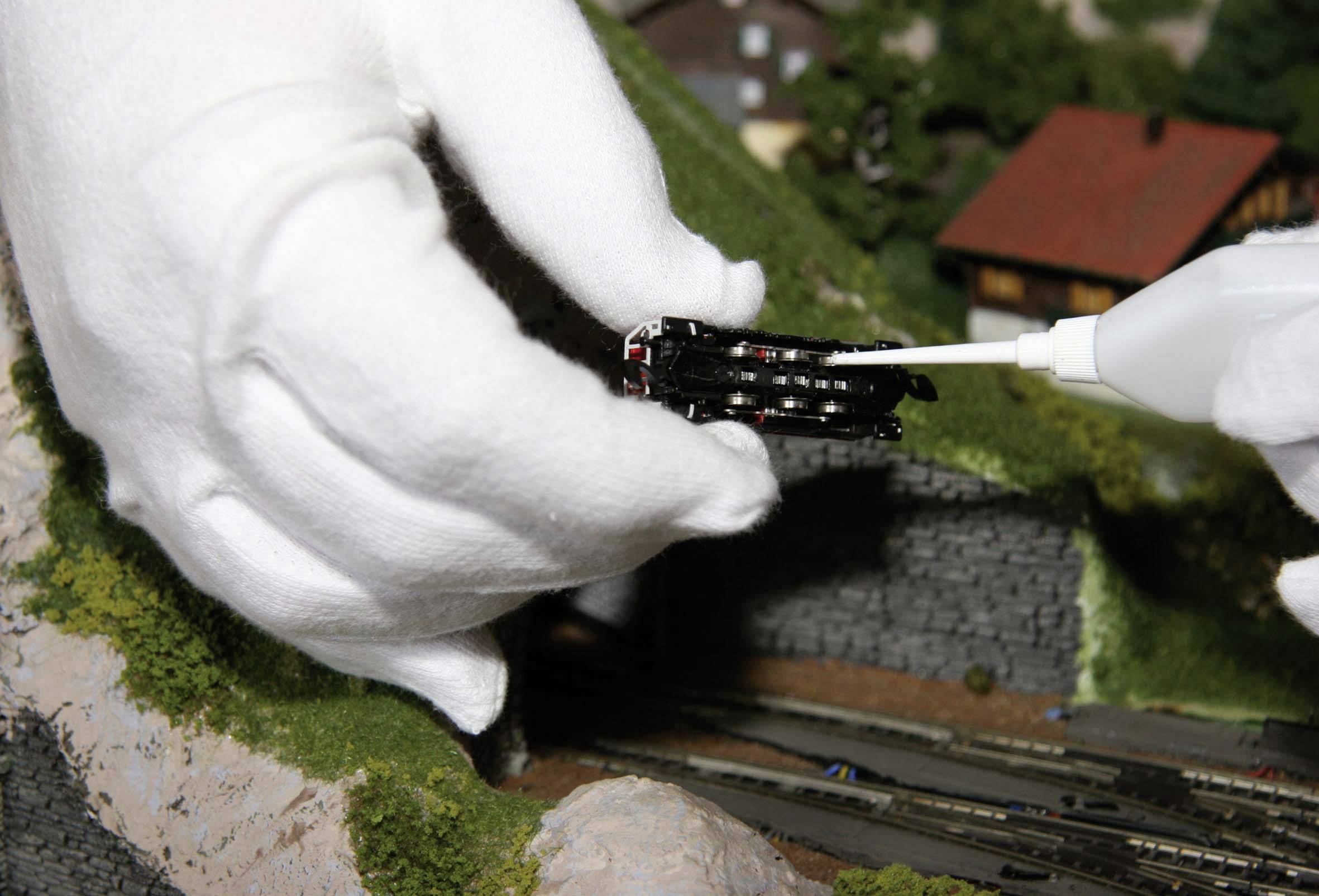 Osoba w białych rękawiczkach przykleja małą miniaturę pociągu klejem w krajobrazie makiety kolejowej.