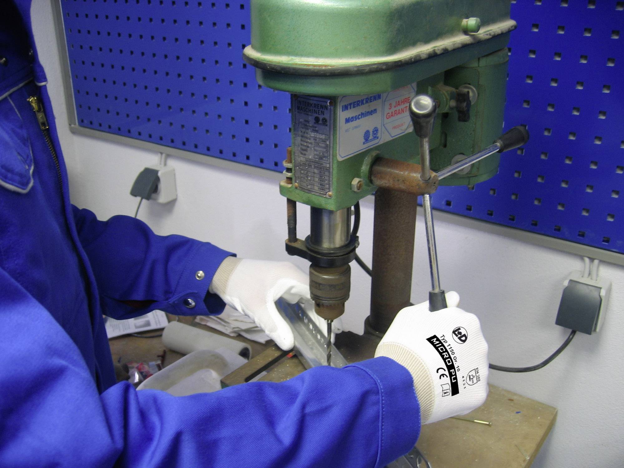 Osoba ubrana w niebieskie ubranie robocze używa stacjonarnej wiertarki do obróbki kawałka metalu. Ma na rękach białe rękawice robocze.