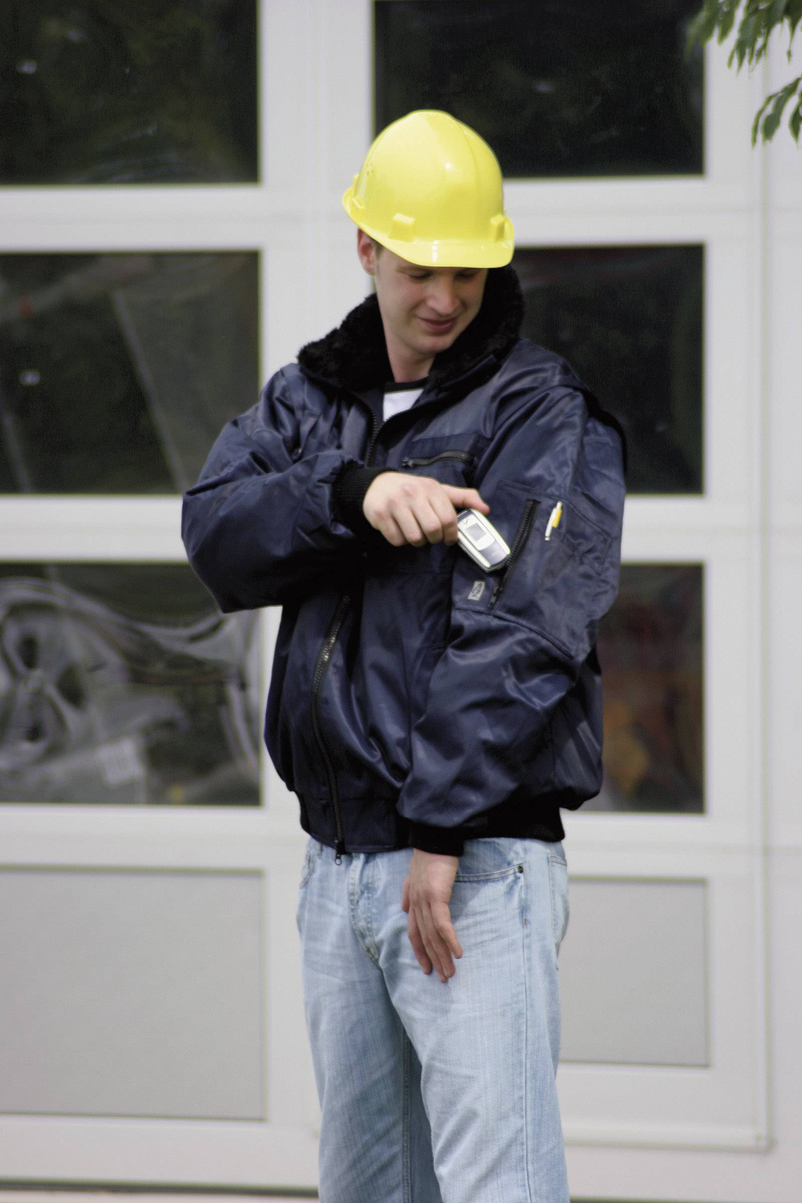 Osoba nosi żółty kask ochronny i ciemną kurtkę. Używa urządzenia na swoim ramieniu. W tle widać bramę garażową.