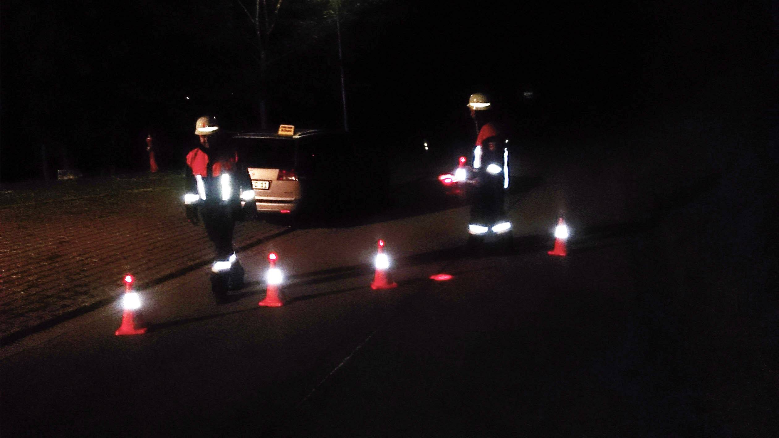 Nocna scena, w której dwie osoby w kamizelkach odblaskowych stoją z latarkami wokół kilku ustawionych na drodze pachołków drogowych.