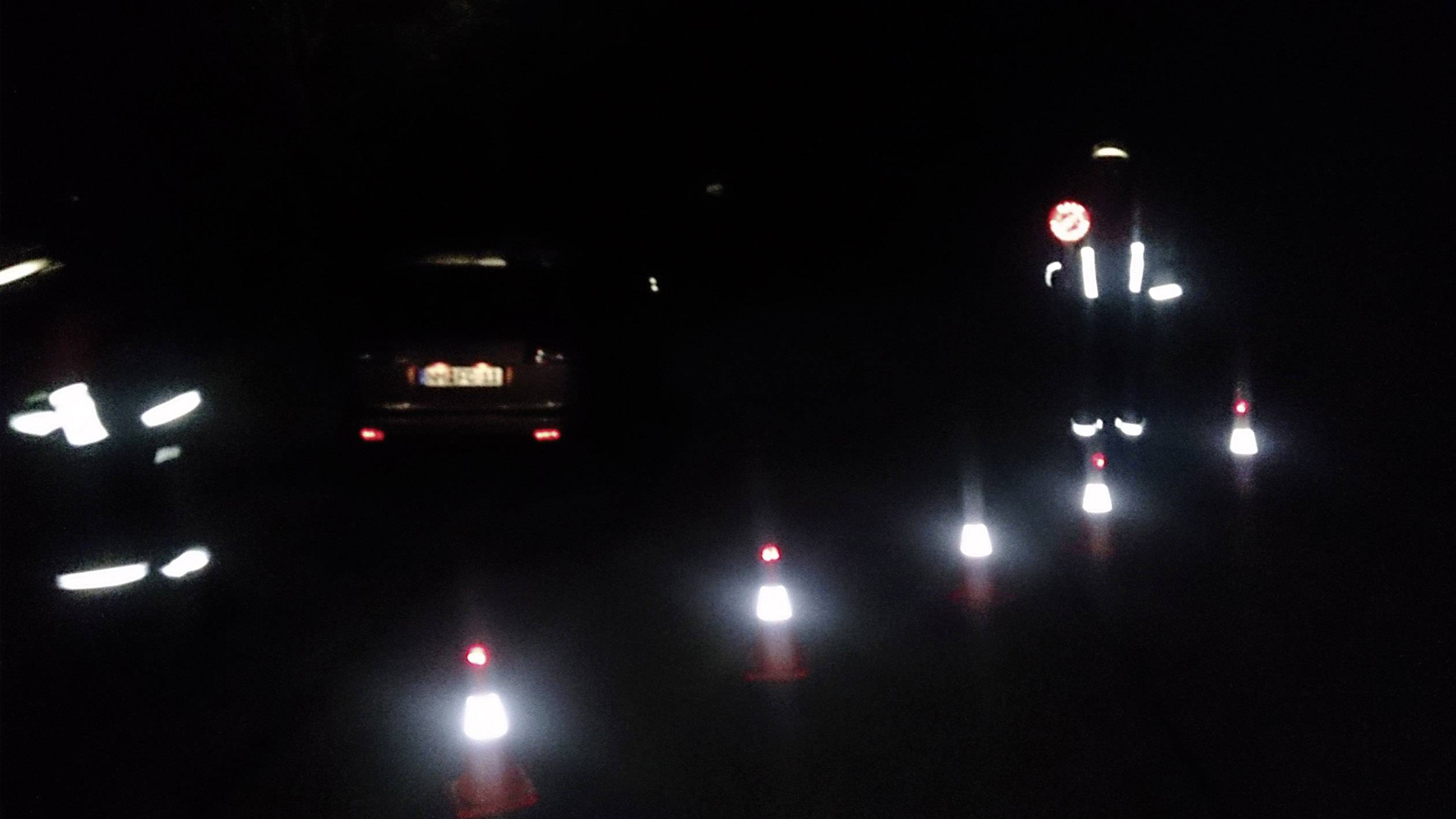 Ciemna ulica jest zamknięta pachołkami drogowymi, podczas gdy osoba w odblaskowym ubraniu stoi obok zaparkowanego samochodu.