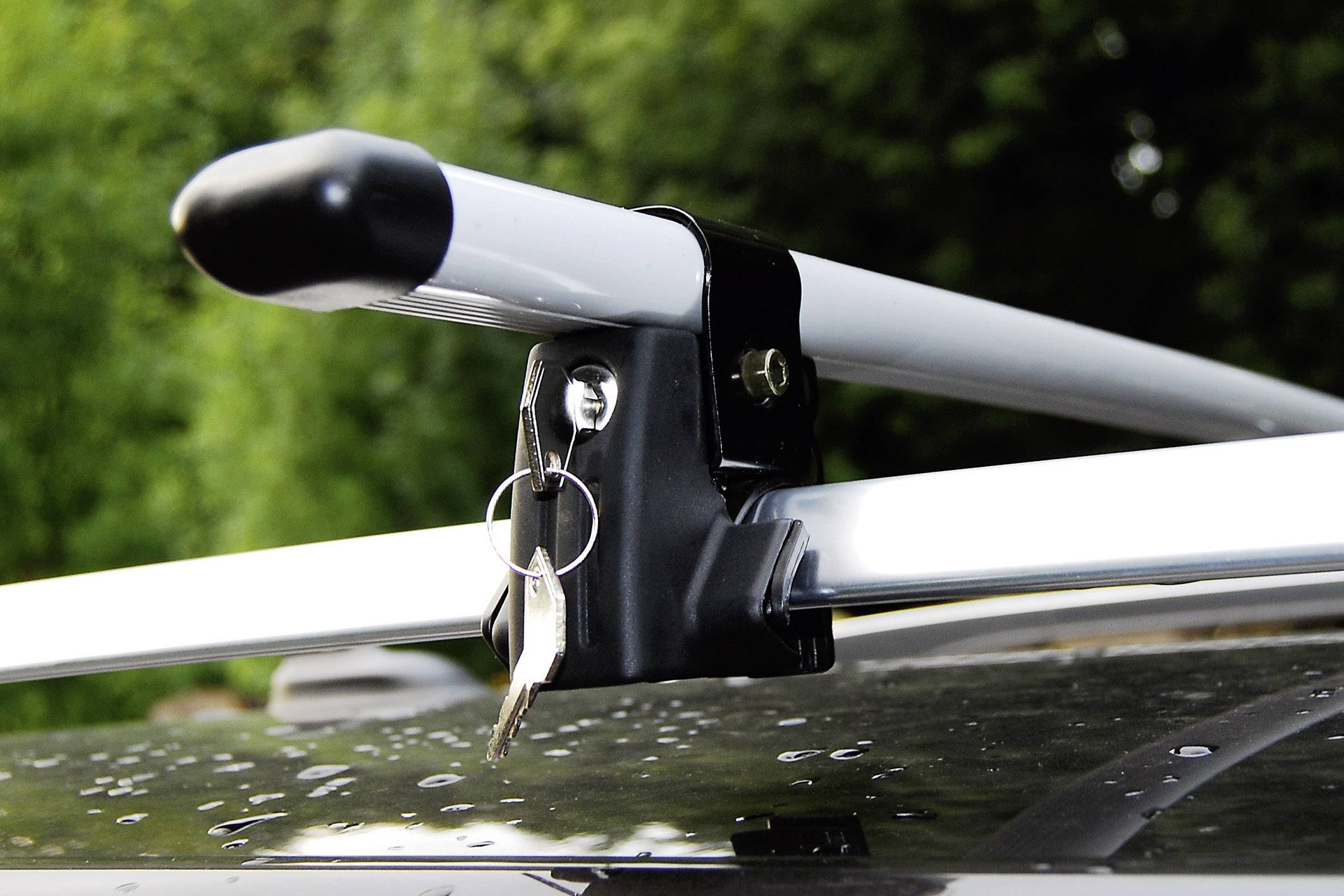 Bagażnik dachowy na samochodzie z włożonym kluczykiem, widoczny na mokrej powierzchni, otoczony zieloną roślinnością w tle.