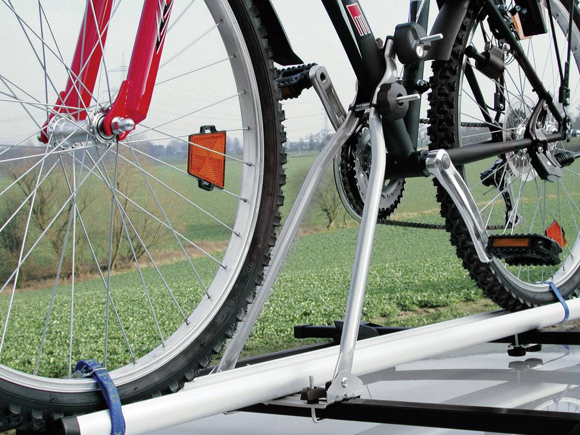 Dwa rowery na bagażniku dachowym samochodu, przymocowane uchwytami. W tle widać zielone pola i pochmurne niebo.