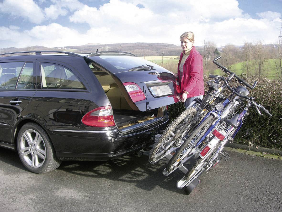 Kobieta stoi obok samochodu z bagażnikiem rowerowym, na którym zamocowanych jest kilka rowerów. W tle widać krajobraz.