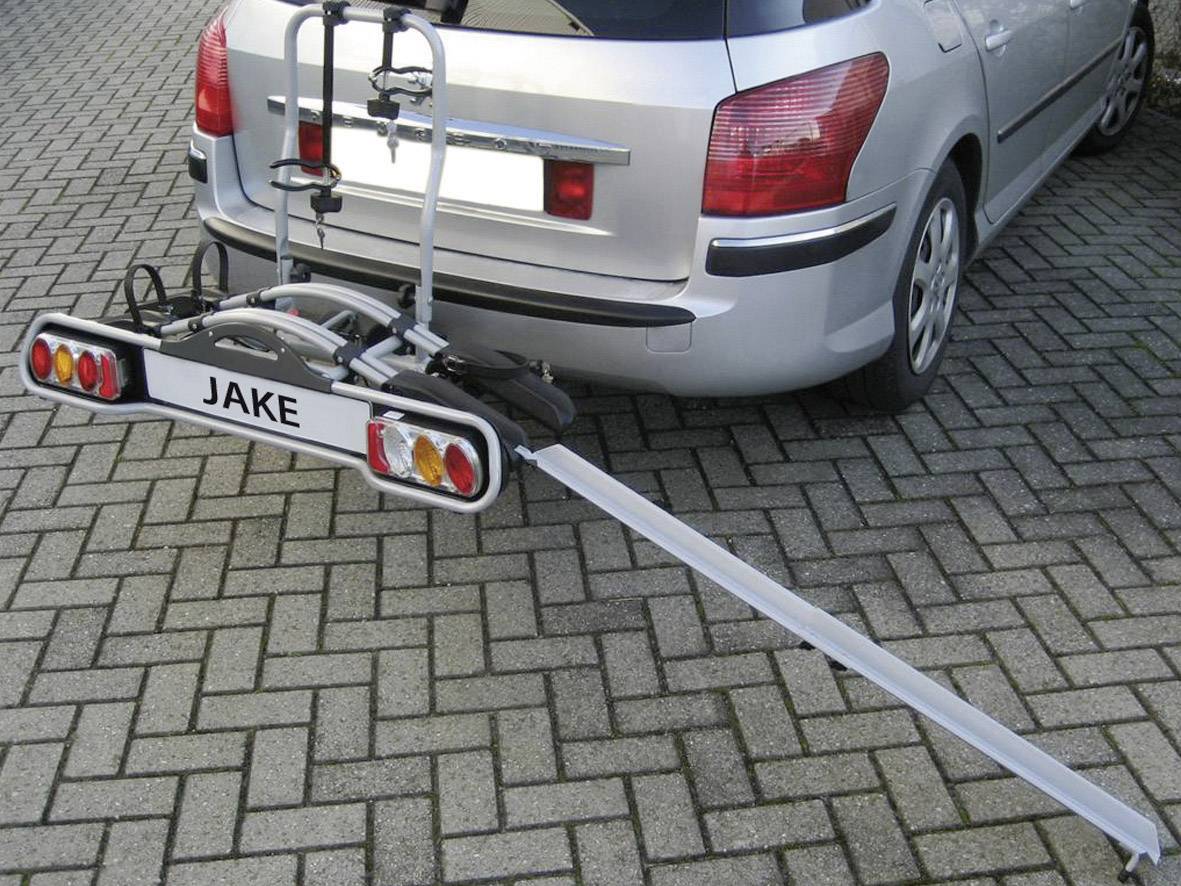 Srebrny samochód z bagażnikiem rowerowym z tyłu, stojący na wybrukowanym parkingu. Na bagażniku widnieje tabliczka z napisem 'JAKE'.