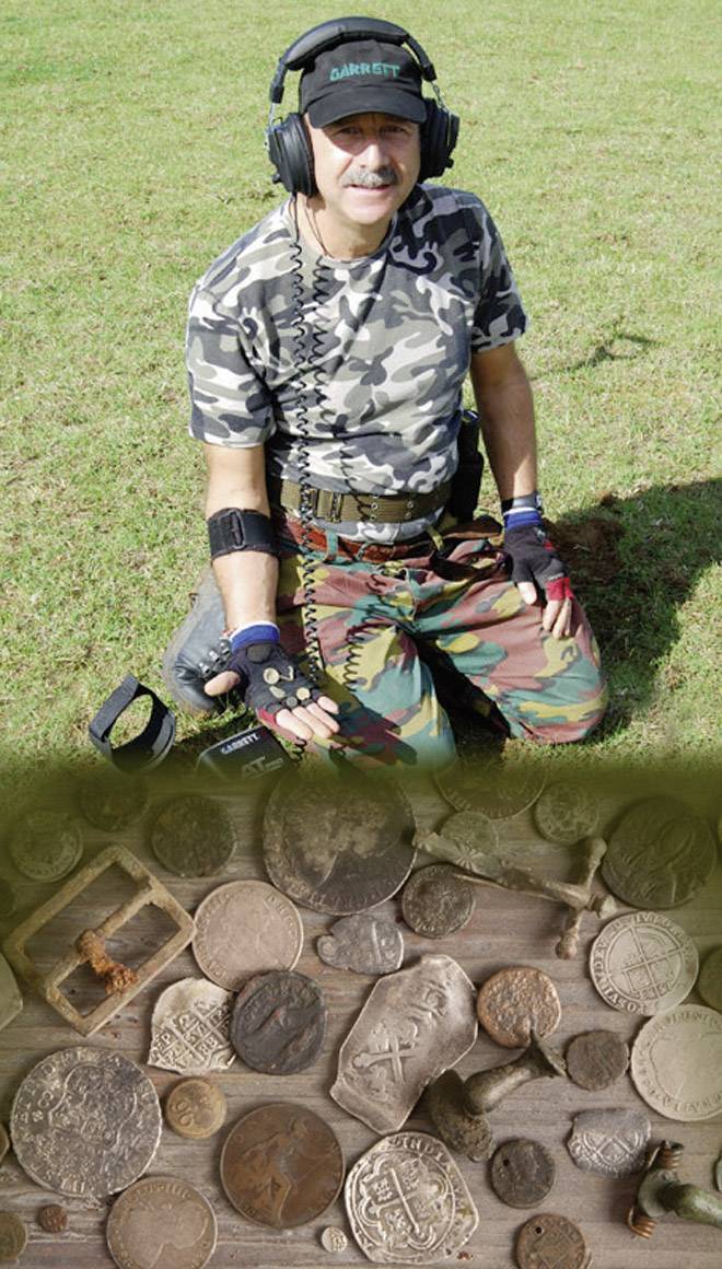 Osoba klęczy na trawie z wykrywaczem metalu, ubrana w odzież maskującą. Na pierwszym planie widoczna jest kolekcja znalezionych monet i artefaktów.