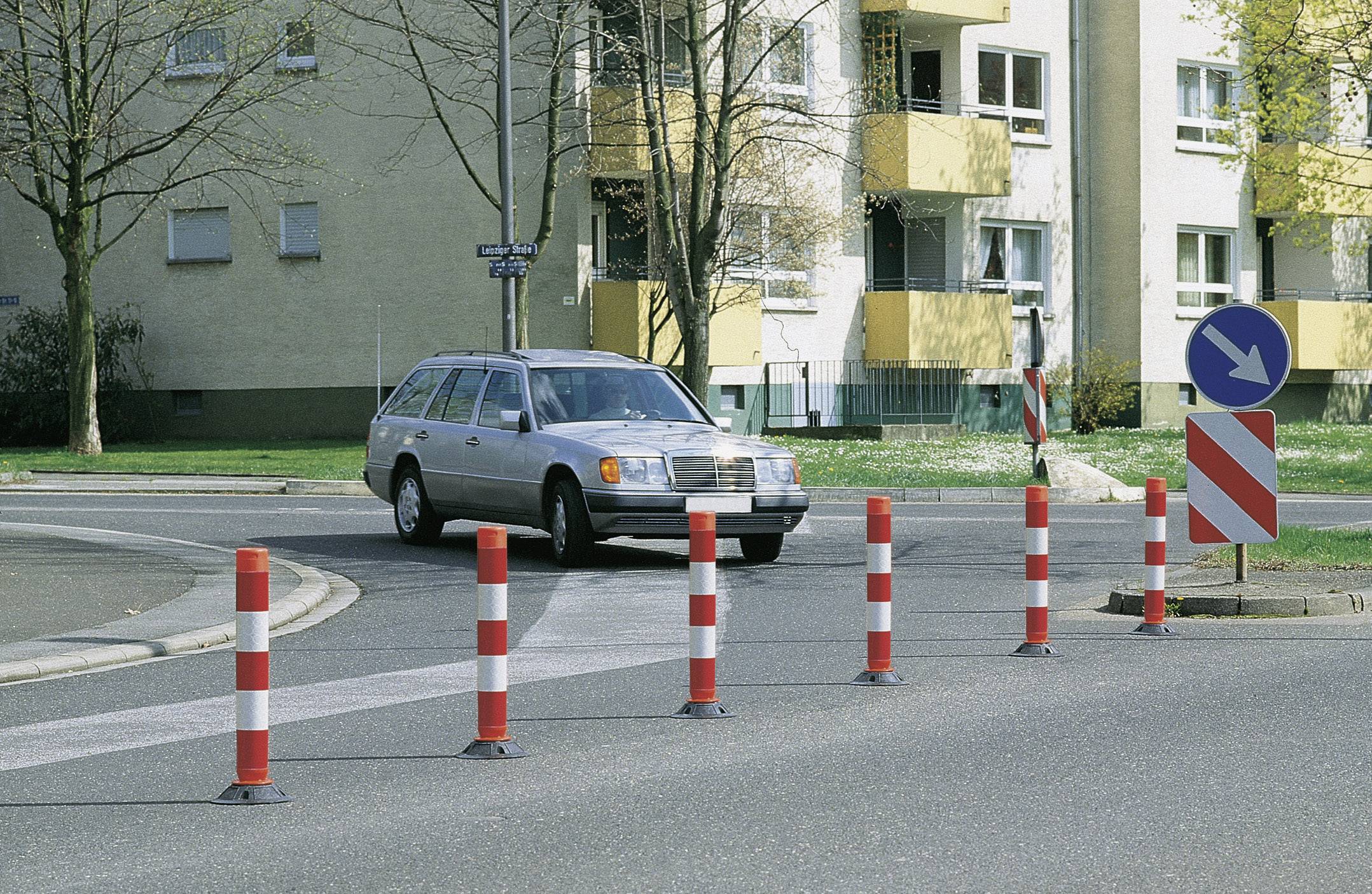 Srebrny samochód przejeżdża przez rondo. Czerwone i białe słupki kierunkowe prowadzą ruch. W tle widać budynki mieszkalne.