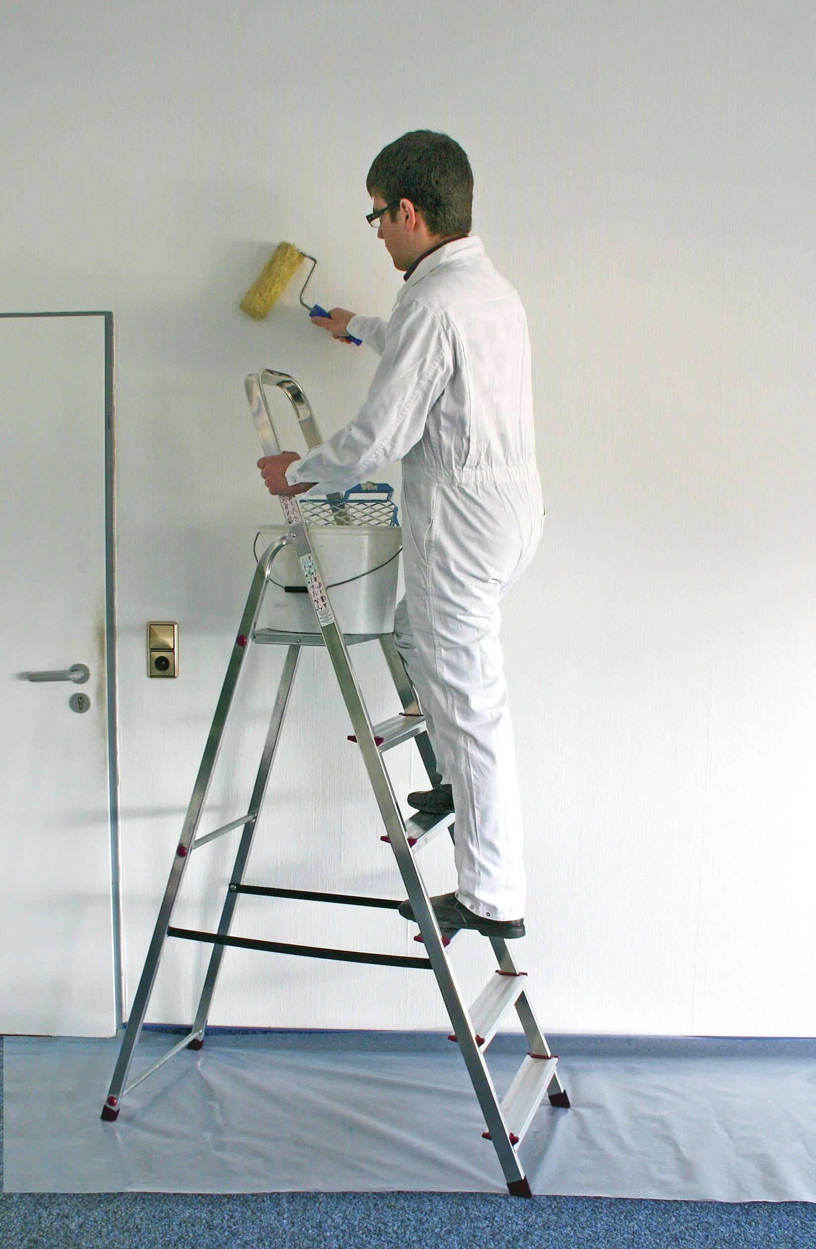 Osoba w białym kombinezonie stoi na drabinie i wałkiem malarskim maluje ścianę na biało.