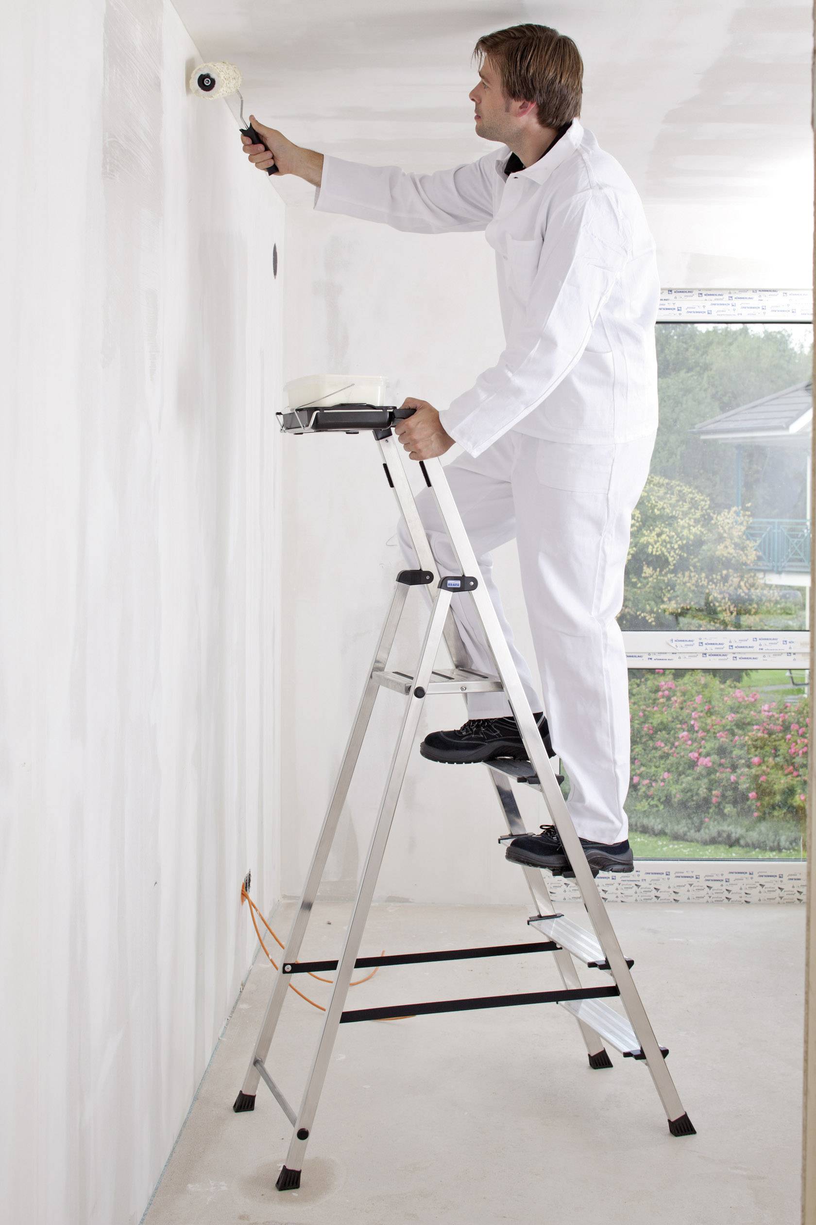 Malarz w białym ubraniu roboczym stoi na drabinie i maluje białą ścianę wałkiem. W tle widać okno.