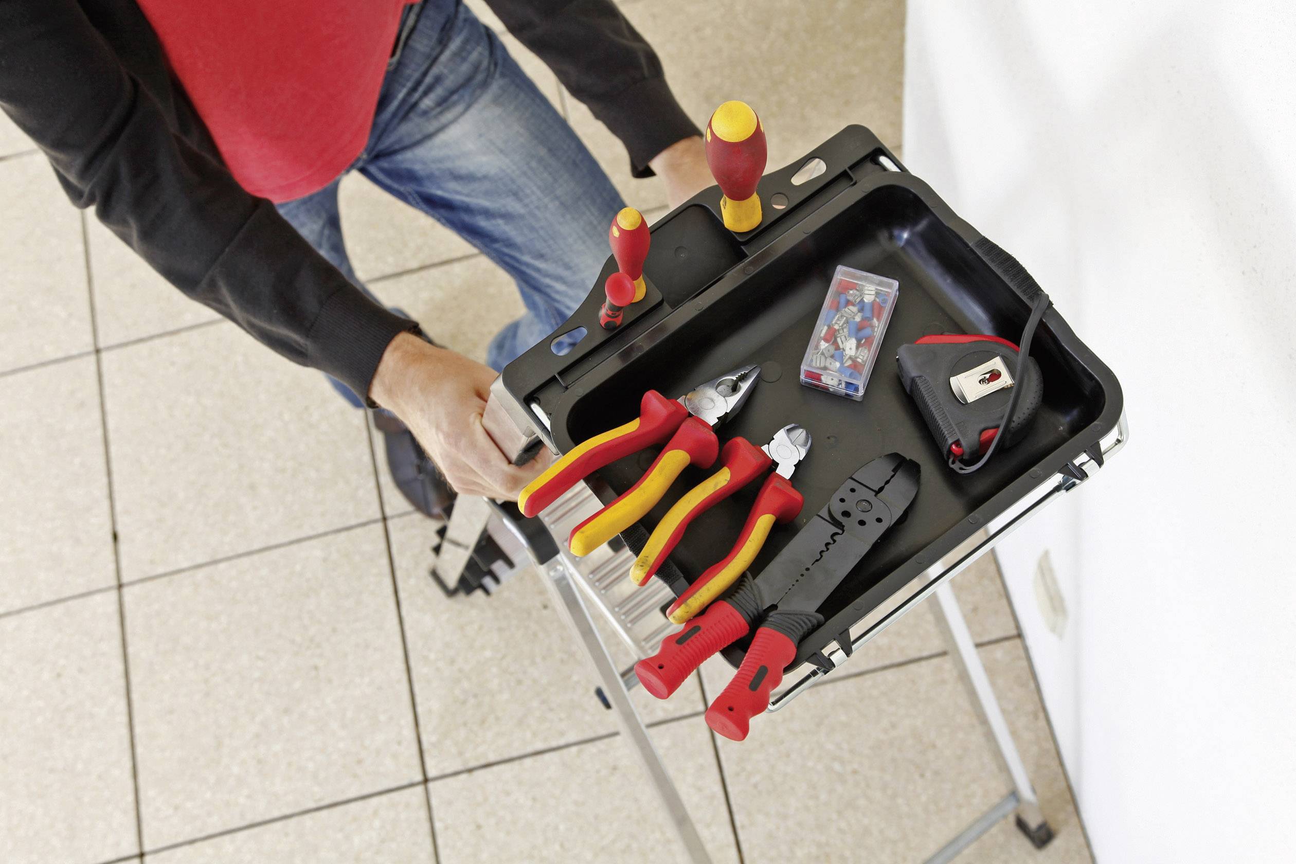 Osoba nosi czarną skrzynkę narzędziową z wkrętakami, szczypcami, taśmą mierniczą i pudełkiem na drobne elementy. Osoba stoi na drabinie.
