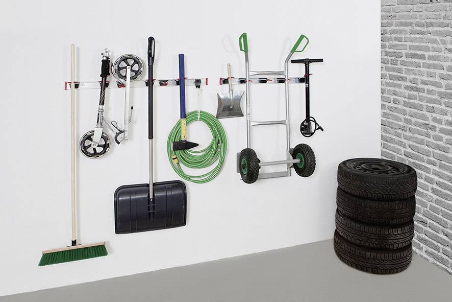 Narzędzia: miotła, łopata, wąż i taczka, starannie powieszone na uchwytach ściennych. Obok stos opon.