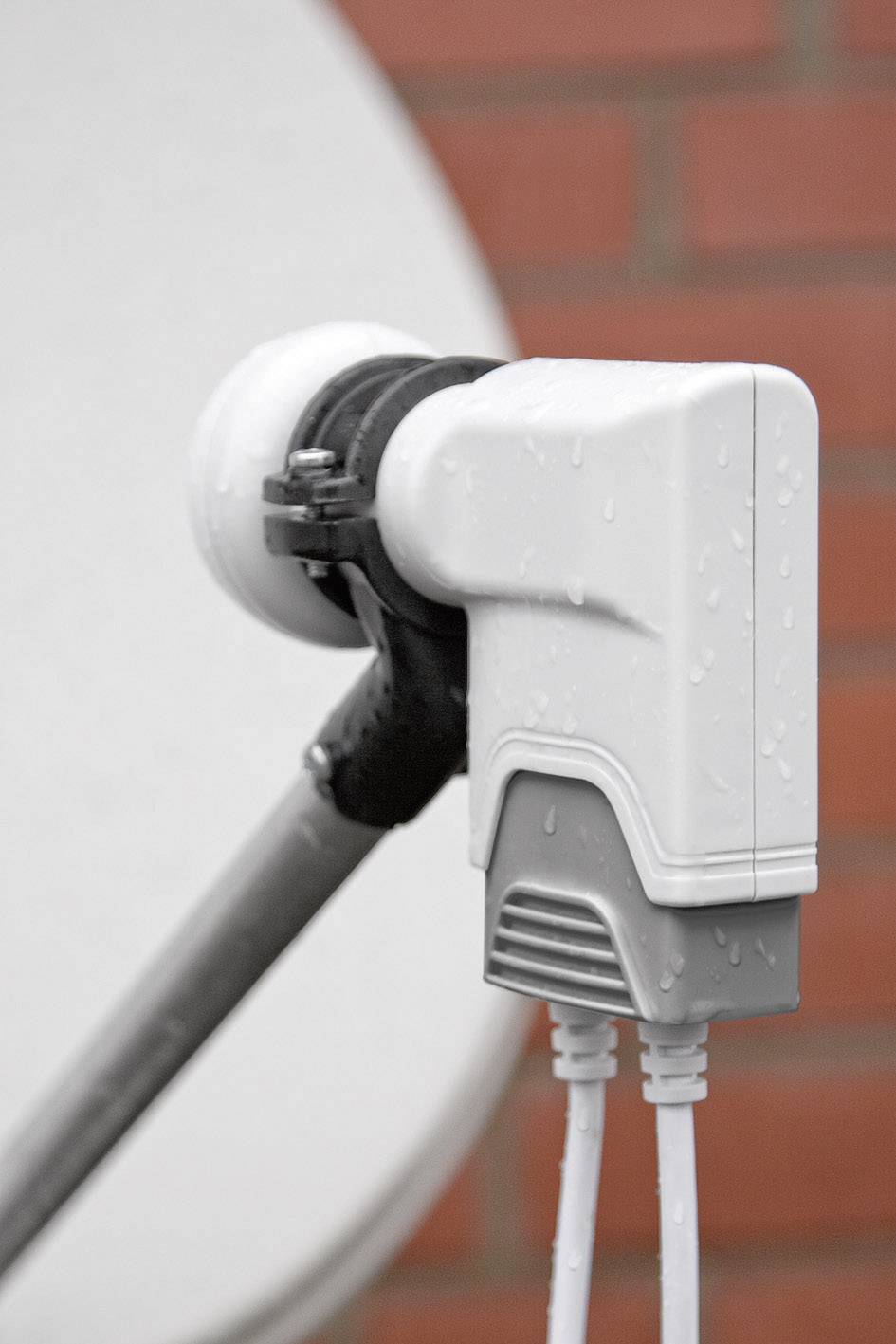 Zbliżenie na konwerter LNB anteny satelitarnej pokryty kroplami deszczu, na tle czerwonego muru z cegły.