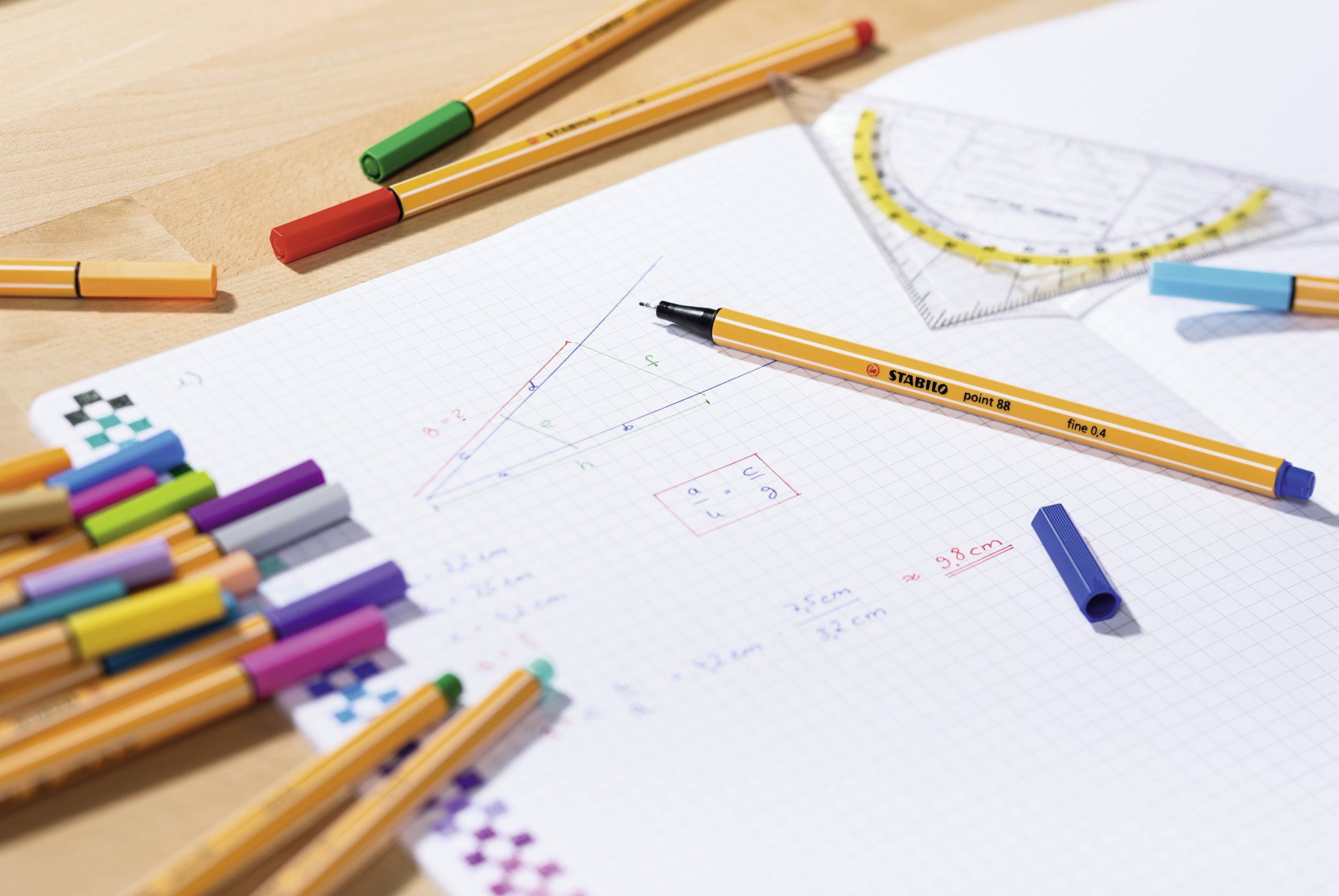 Matematický pracovný list s geometrickými nákresmi, farebnými perami a uhlomerom. Hlavný fokus je na nakreslenom trojuholníku.