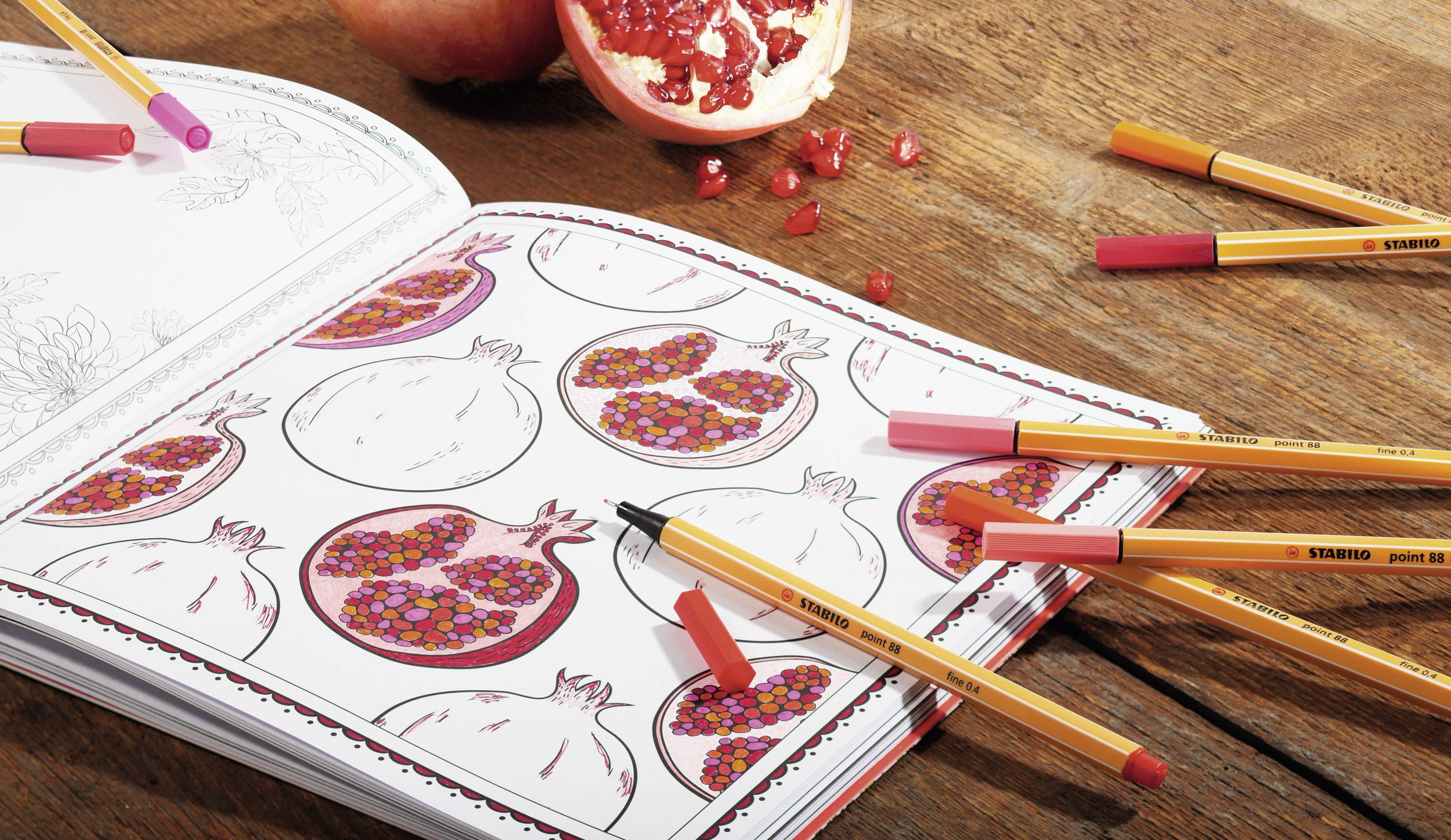 Omaľovánka s otvorenými ilustráciami granátového jablka, obklopená farebnými pastelkami. Vedľa leží rozrezaný granátový plod.