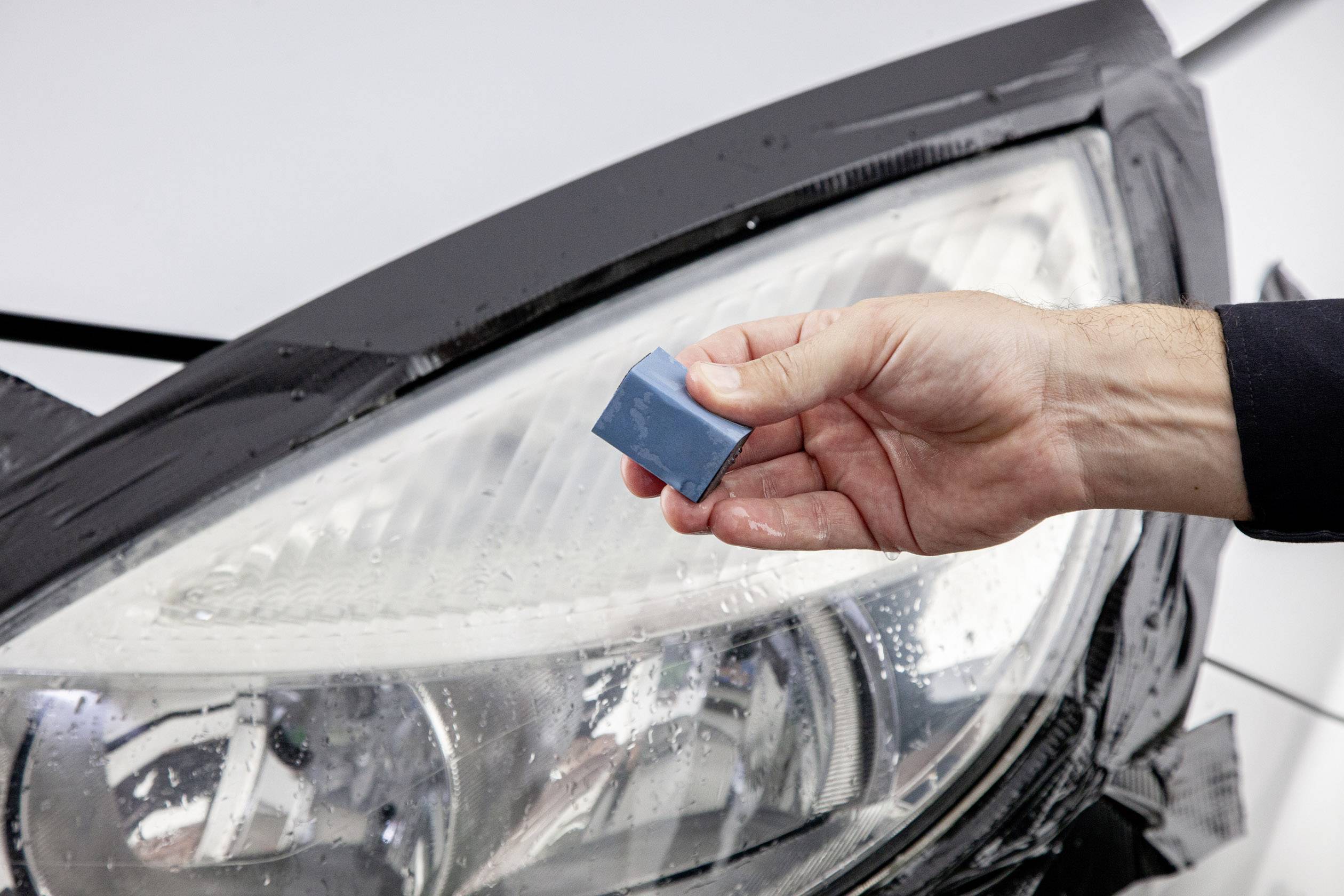 Ruka drží malý modrý predmet pred automobilovým svetlometom pokrytým vodou.