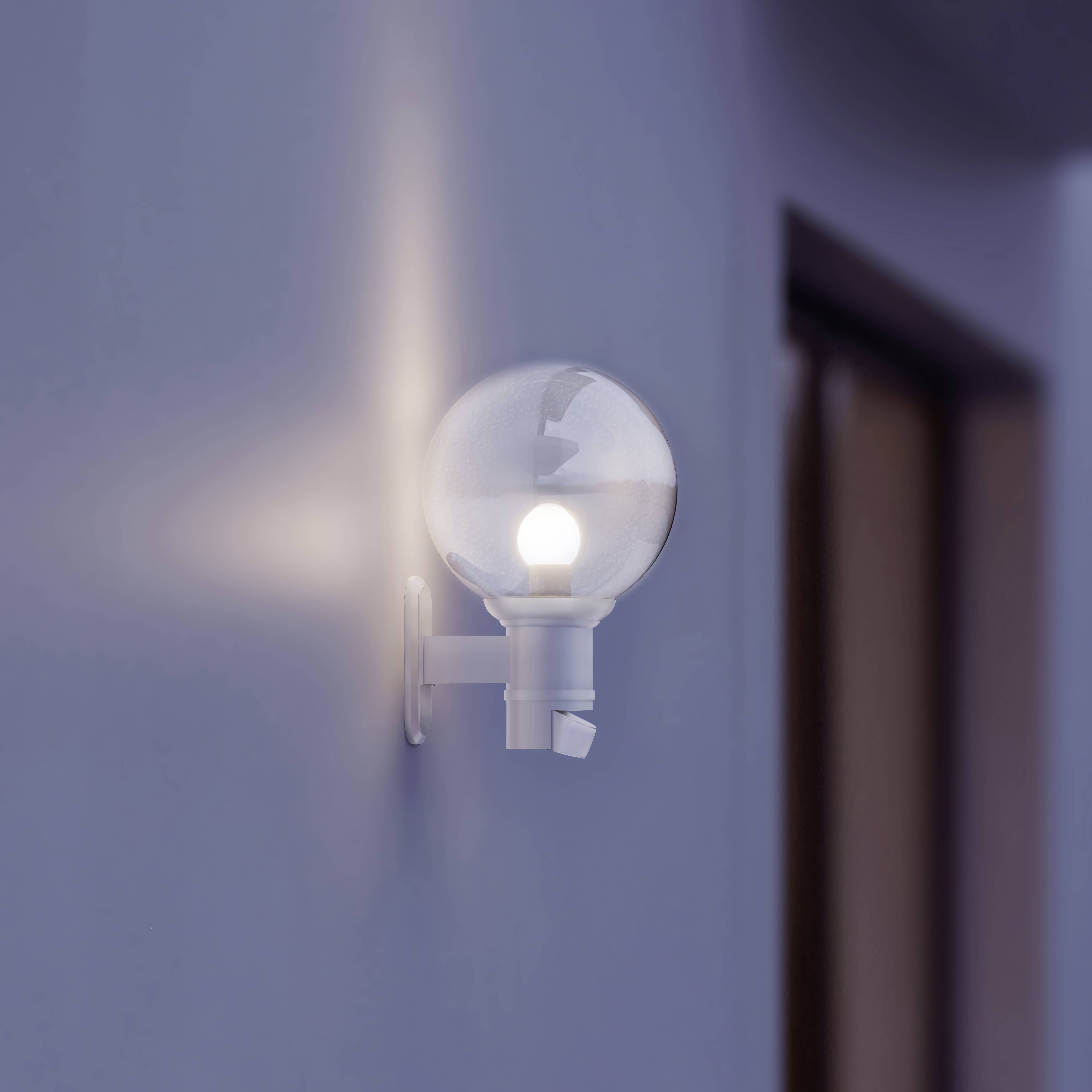 Moderná nástenná lampa s guľatou sklenenou guľou svieti oproti modrej stene a jemne osvetľuje priľahlý priestor.