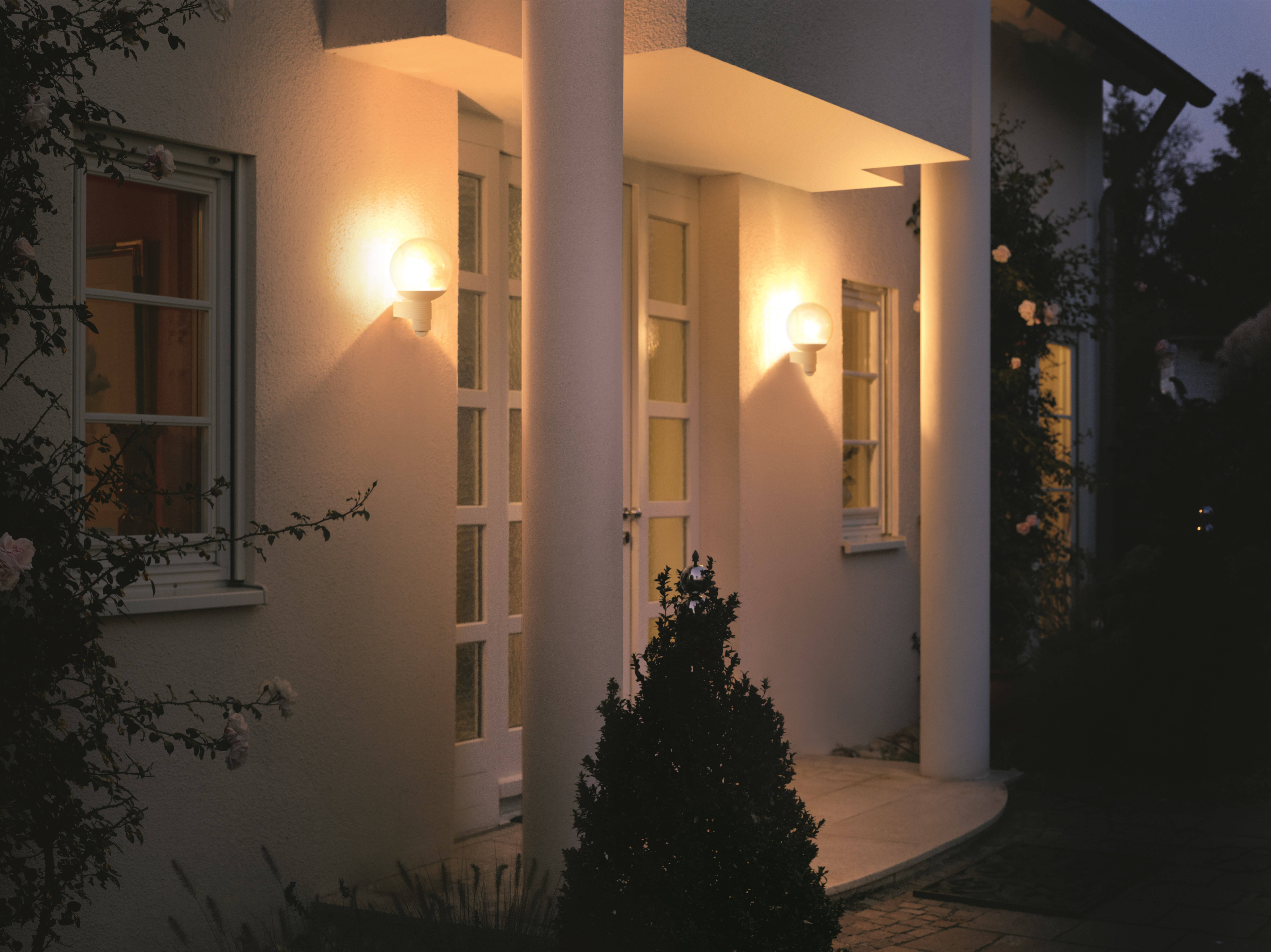 Vstup domu za súmraku, osvetlený dvoma vonkajšími stenými svietidlami. Viditeľný je stĺp a kríky.