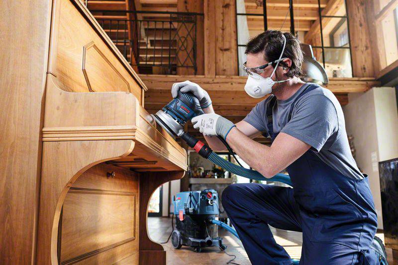 Muž nosí ochrannú masku a rukavice, pričom obrába drevený stôl elektrickou brúskou v miestnosti.