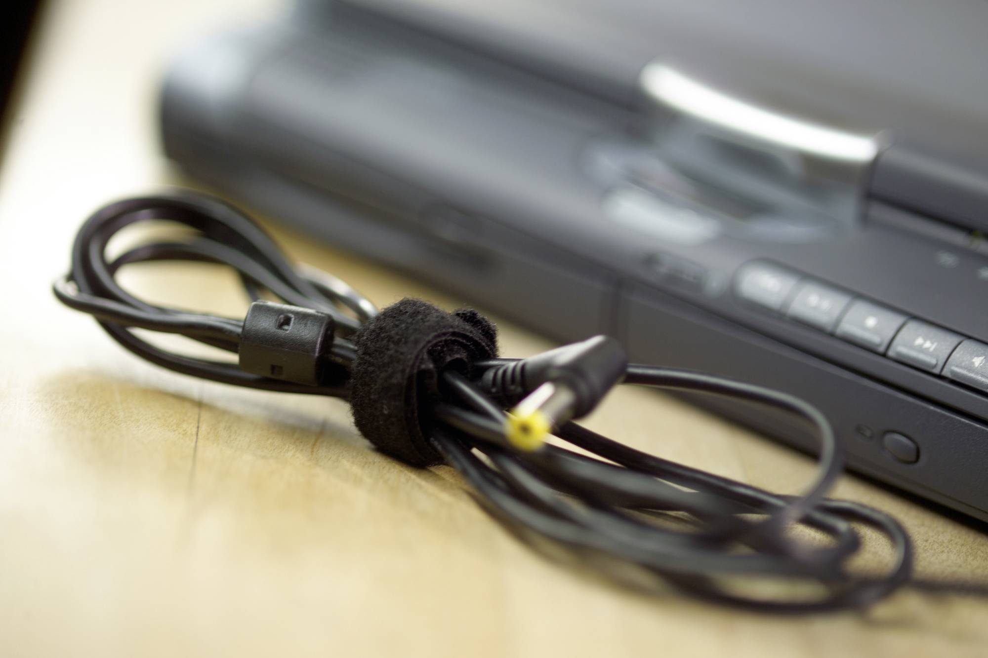 Na drevenom stole leží zvinutý sieťový kábel pred rozmazaným elektronickým zariadením v pozadí.