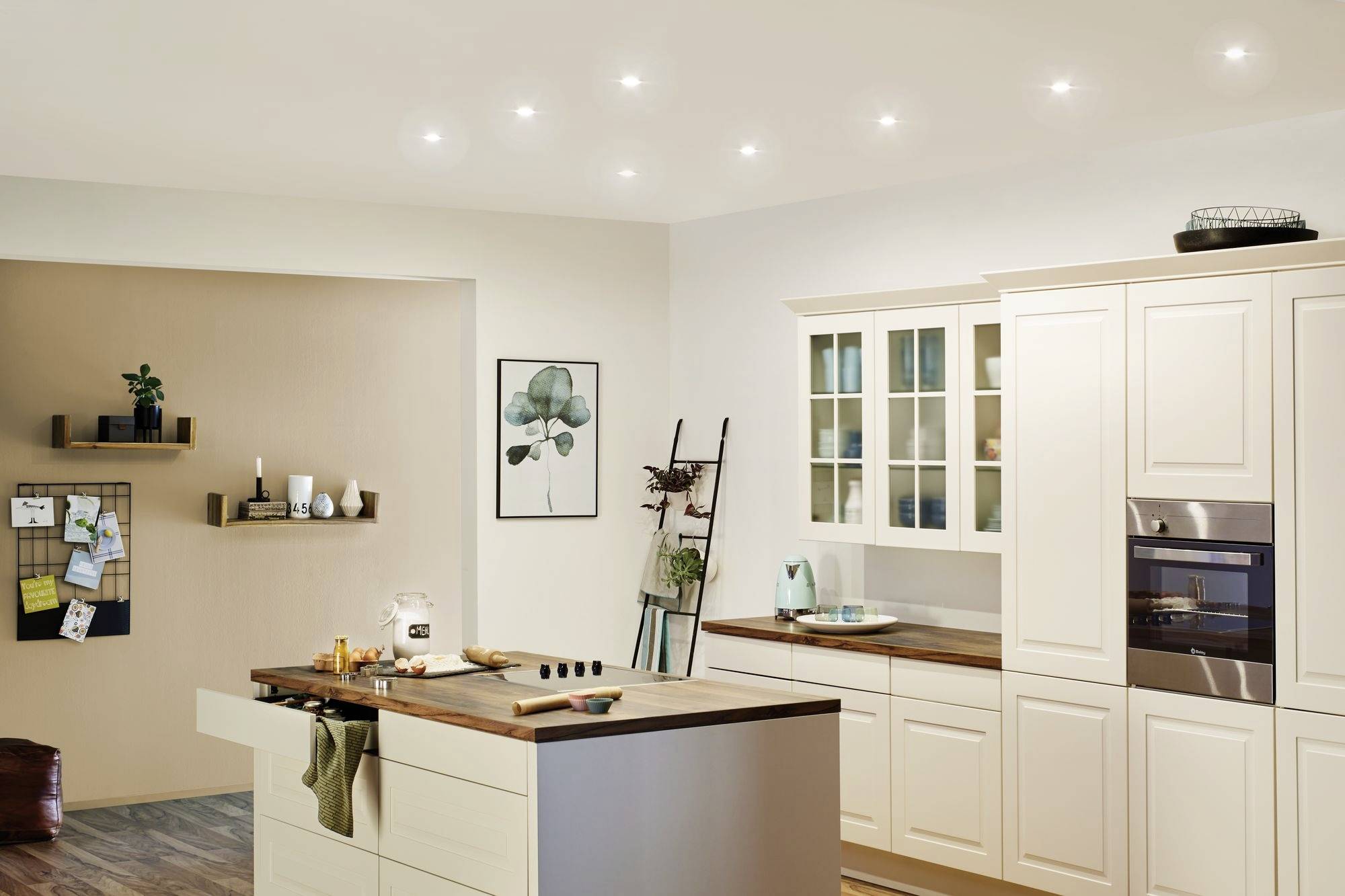 Svetlá moderná kuchyňa so stredovým ostrovčekom, bielymi skrinkami a drevenými pracovnými doskami. Na stene sú zavesené police a dekorácie.