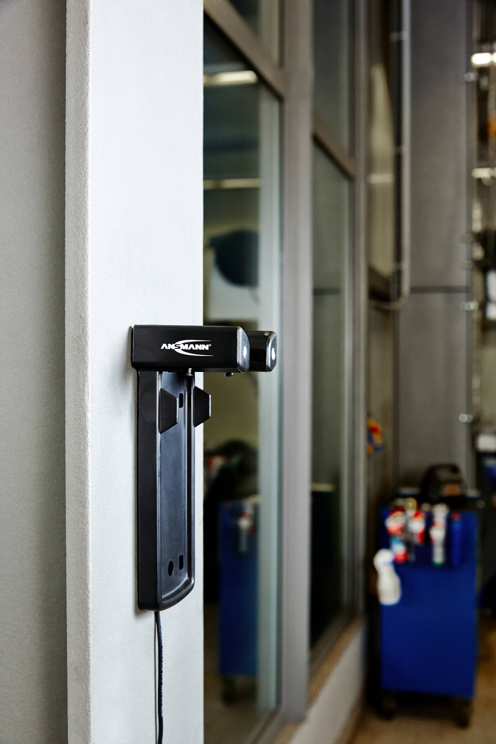 Čierna nabíjacia stanica pre elektrické náradie je namontovaná na stene. V pozadí sa nachádza vozík s rôznymi nástrojmi.