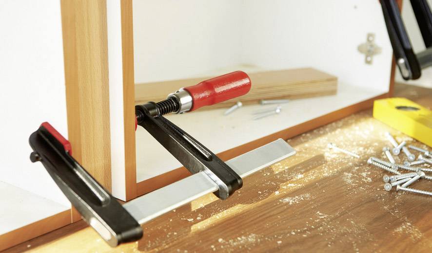 Drevená pracovná lavica so svorkou, ktorá spája dva dosky. Na povrchu ležia skrutky a žltá vodováha.