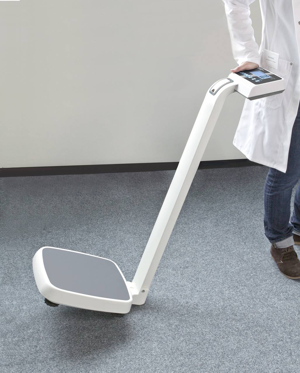 Osoba v bielom plášti drží moderné, prenosné medicínske zariadenie s displejom na sivom koberci.