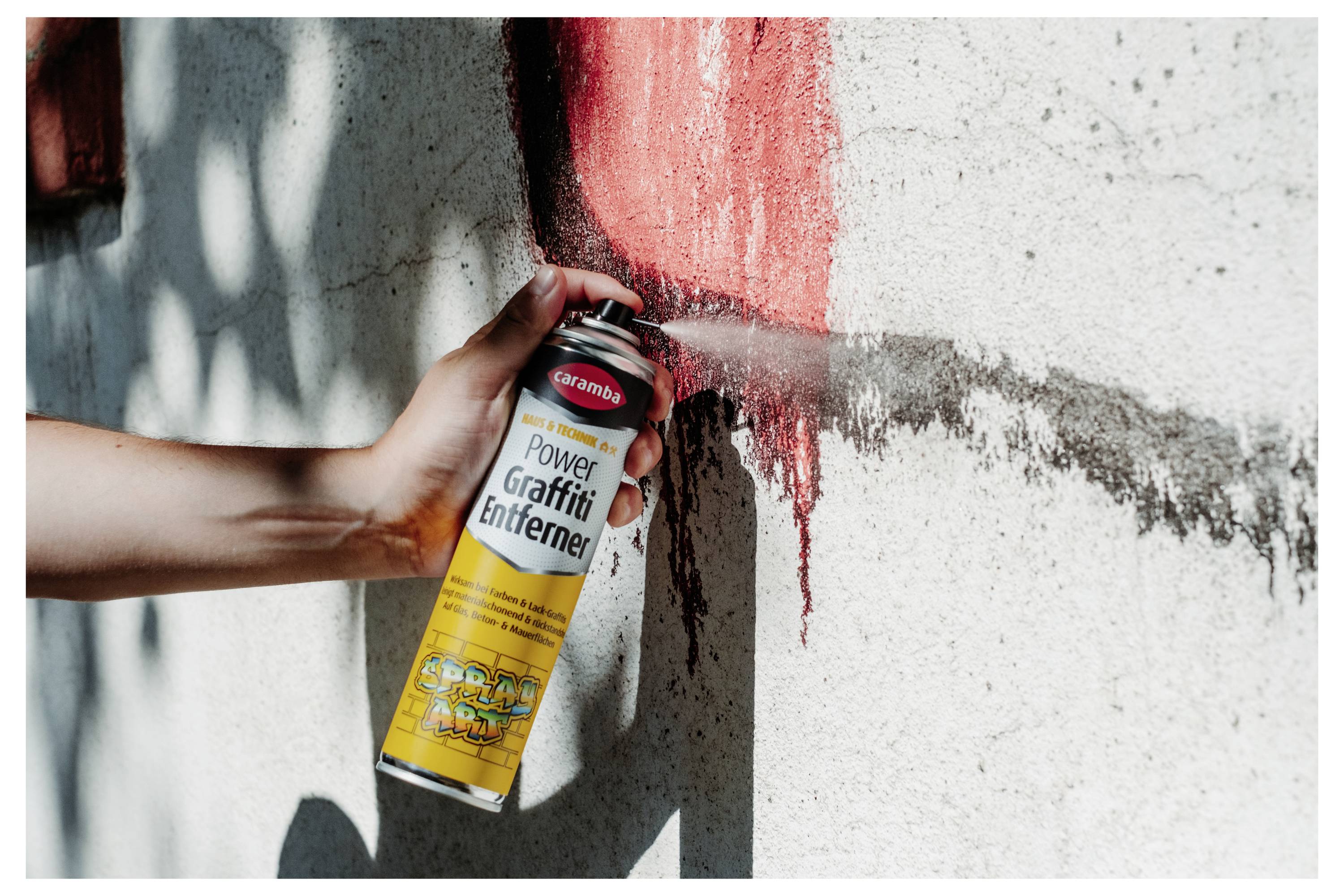 Ruka strieká odstraňovač graffiti z nádoby s nápisom „Power Graffiti Entferner" na červenú farbu na stene a čistí povrch.