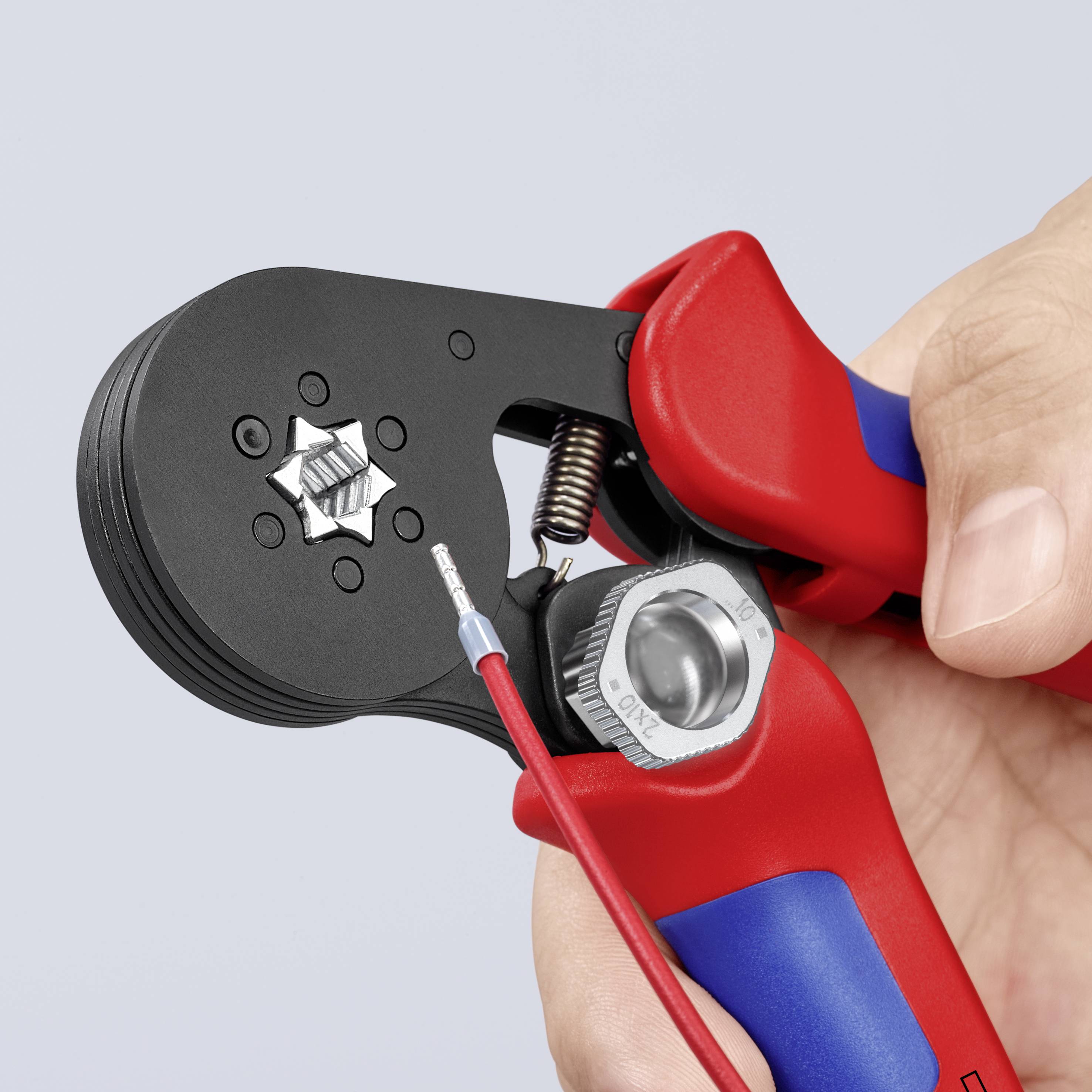 Jedna ruka drží červené krimpávacie kliešte, ktoré spracúvajú drôt s kovovým konektorom.