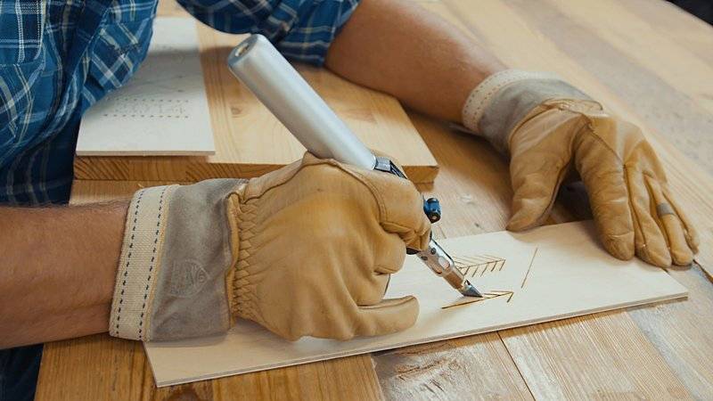 Osoba v rukaviciach vyrýva vzor na drevenej doske pomocou špeciálneho nástroja pri dreveном stole.
