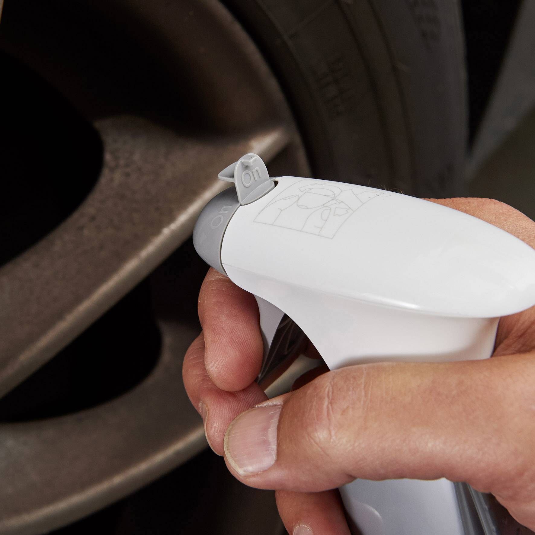 Ruka drží bielu rozprašovaciu fľašu namierenú na ventil pneumatiky automobilu, určenú na čistenie alebo údržbu, pričom je viditeľný kryt ventilu.