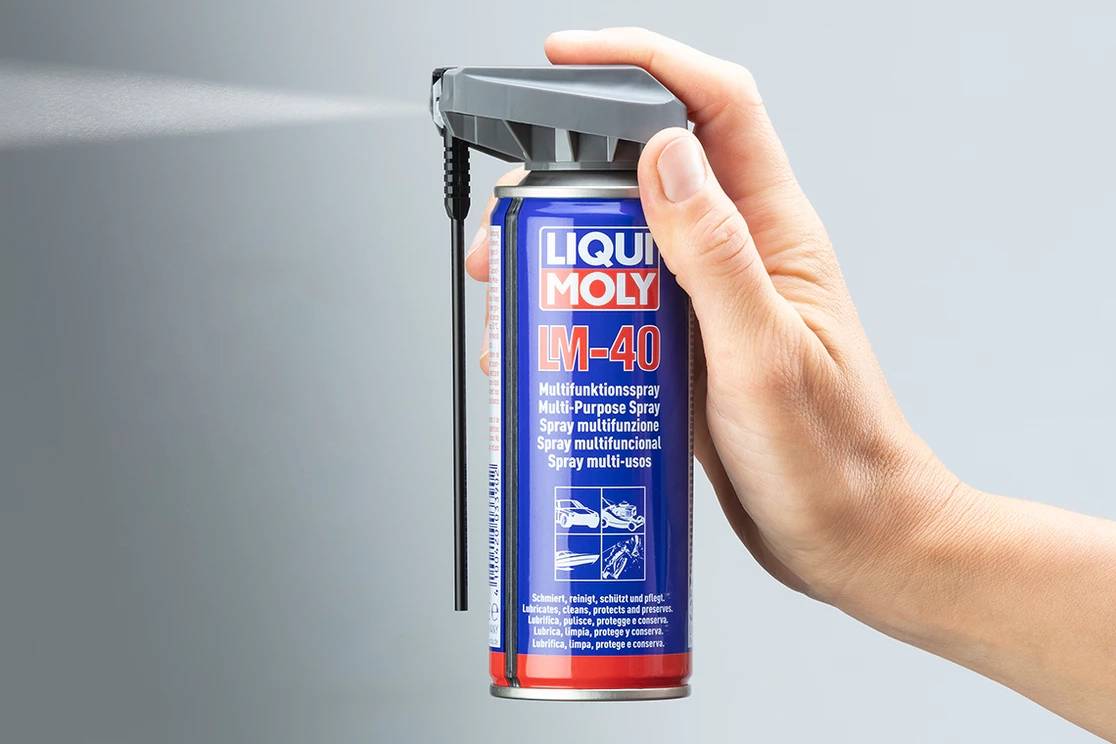 Jedna ruka drží sprejovú konzolu 'Liqui Moly LM-40'. Dóza sa používa; jemná hmla vychádza von.