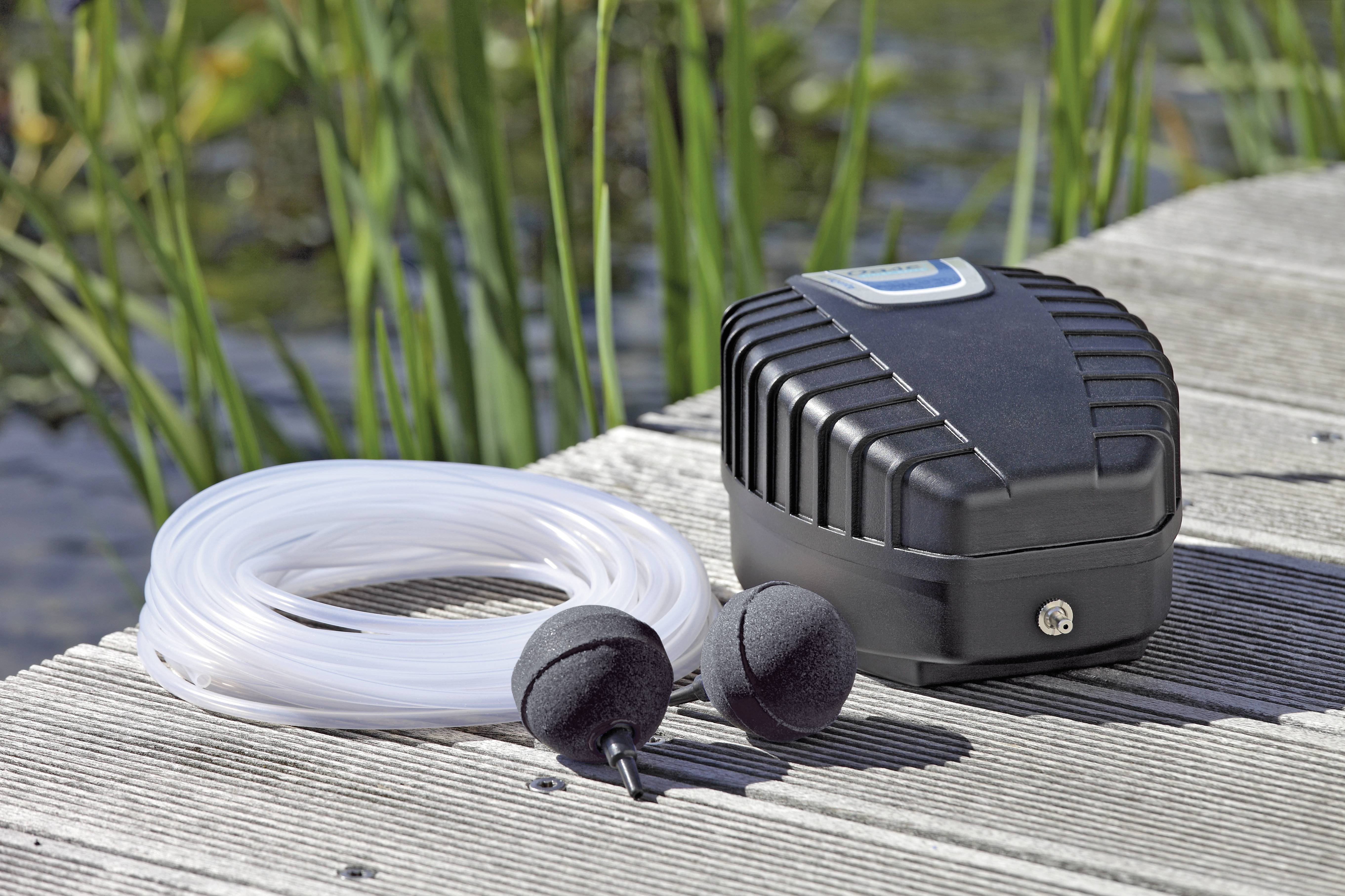 Čierna vzduchová pumpa leží na drevenom móle vedľa zvinutej hadice a dvoch okrúhlych difúzorov, pričom v pozadí sú vysoká tráva a voda.