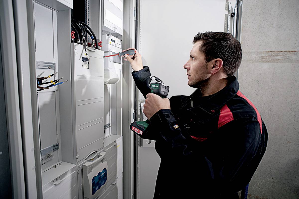 Technik kontroluje elektrickú rozvádzač pomocou skrutkovača a meracieho prístroja. Má na sebe pracovnú bundu.