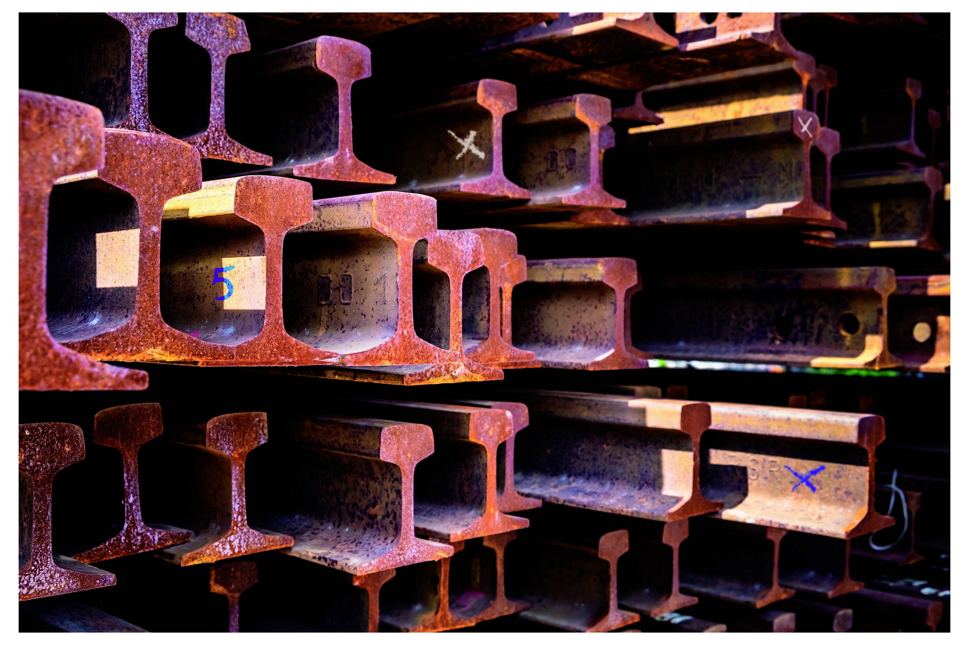 Oceľové I-nosníky uskladnené horizontálne, s viditeľnými farebnými značkami kontroly na niekoľkých nosníkoch.
