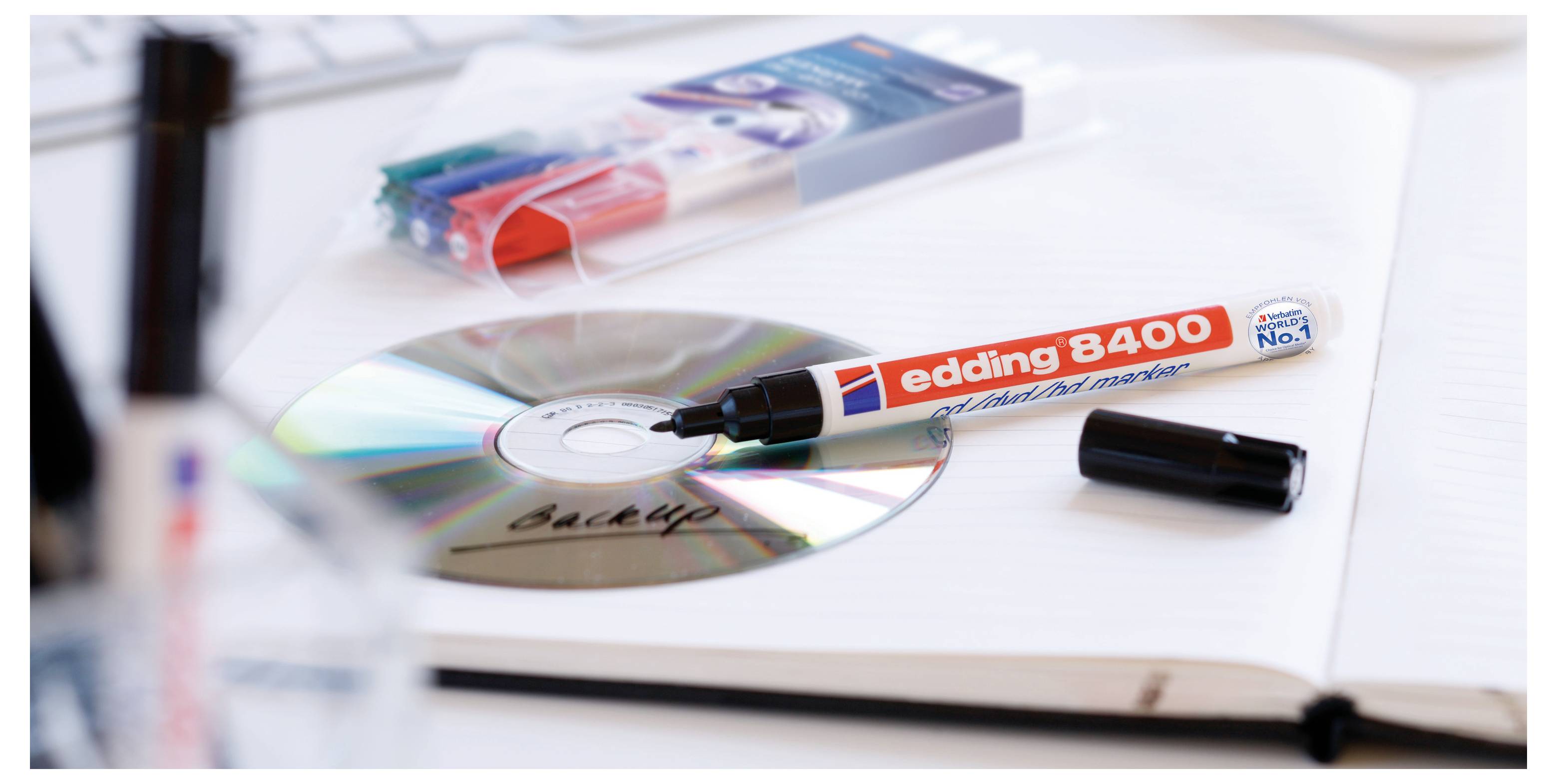 Červeno-čierny značkovací pero „edding 8400" piše na CD vedľa svojho uzáveru a balenia, na bielom zošite alebo pracovnej ploche.