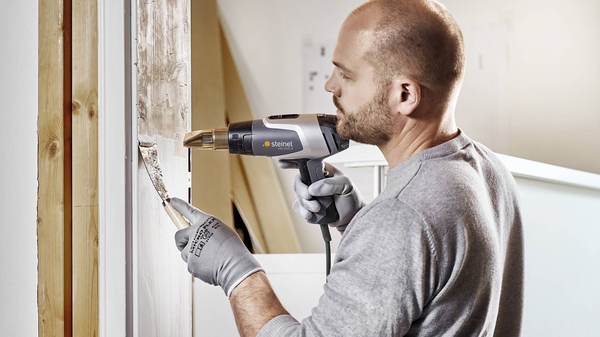 Muž v rukaviciach používa teplovzdušný ventilátor na odstránenie farby z okenného rámu.